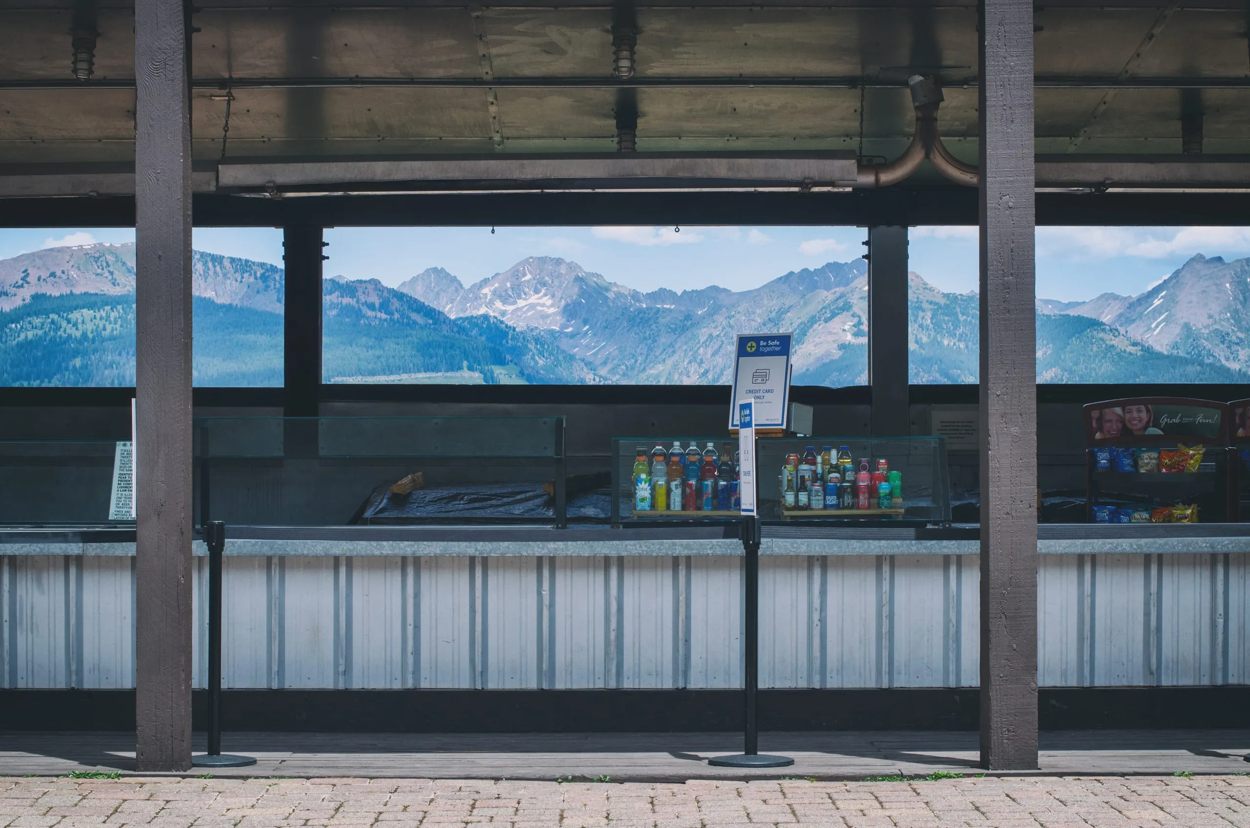 An empty mountaintop concession counter with bottled drinks frames a snow-streaked Colorado mountain range through open windows.