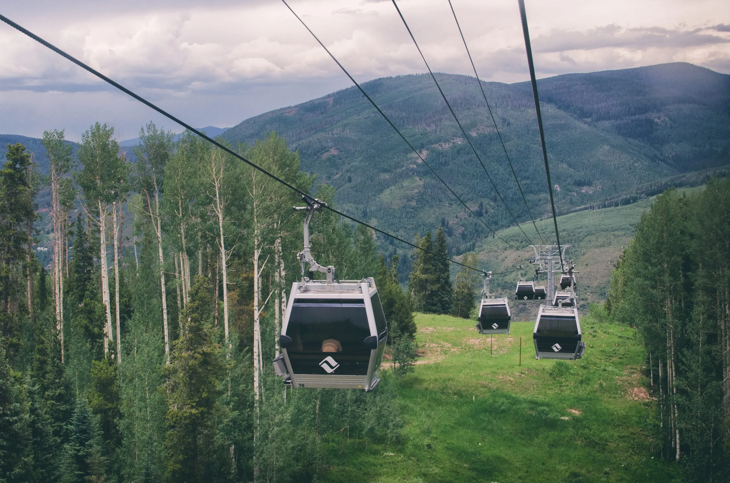Gondola cabins hang from cables above green forested slopes and distant mountains at a Colorado resort.