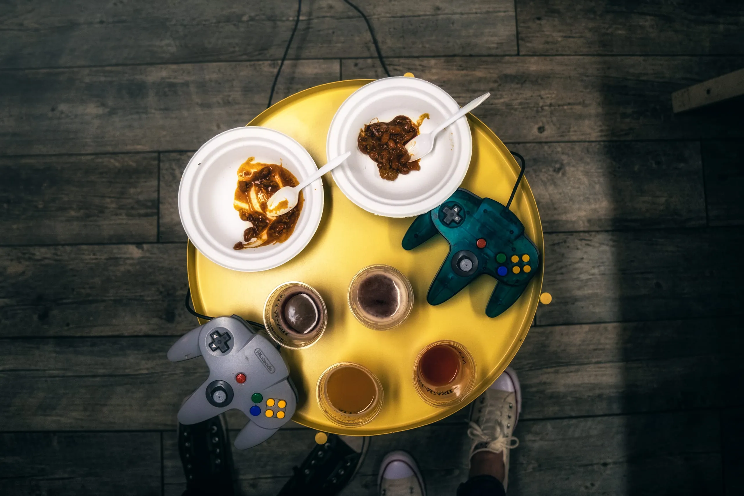 Overhead view of a yellow table with two N64 controllers, a beer flight, and paper plates of chili in Saratoga Springs.