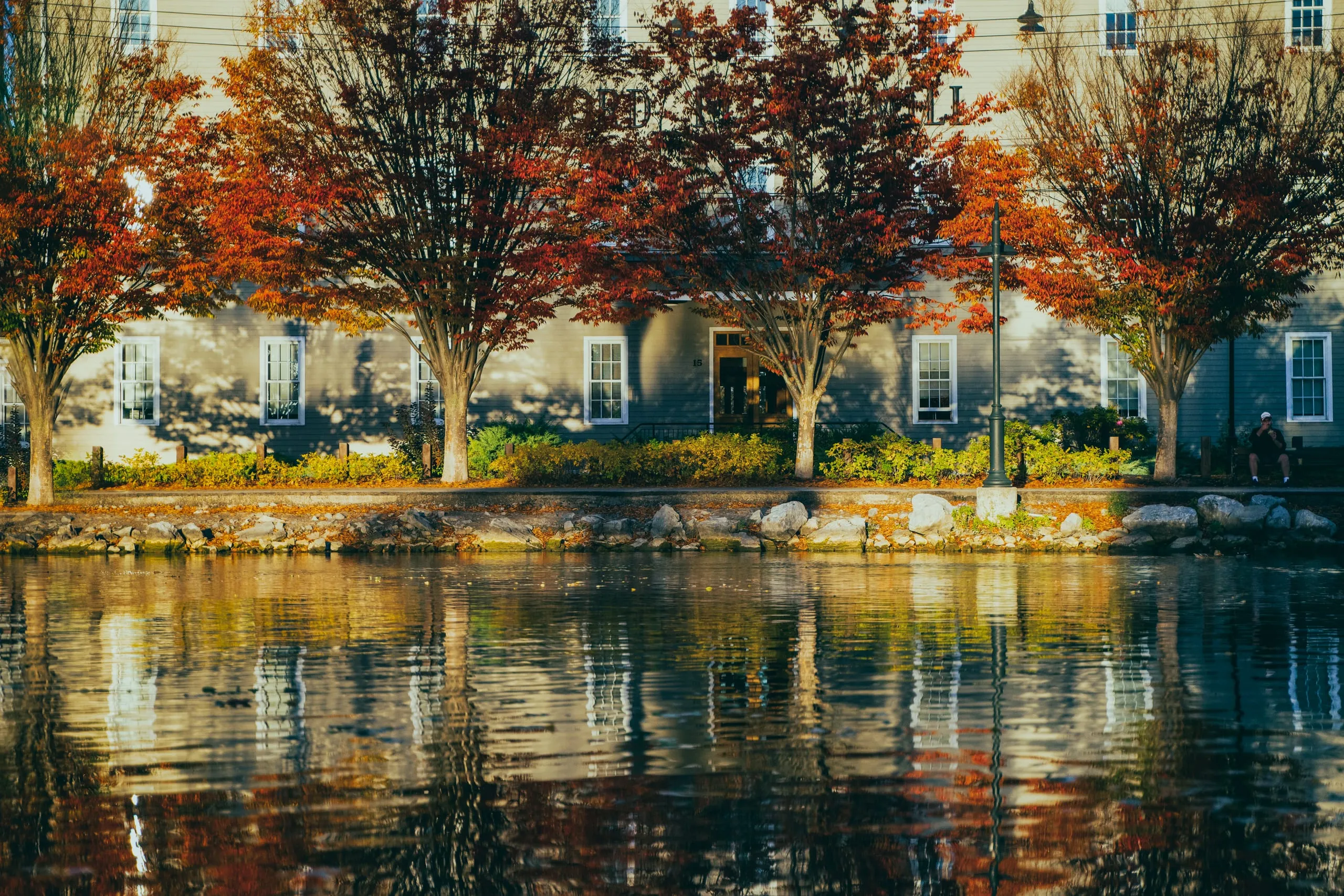 Red-leafed maple trees line a gray clapboard building mirrored in a still canal in Rochester, New York.