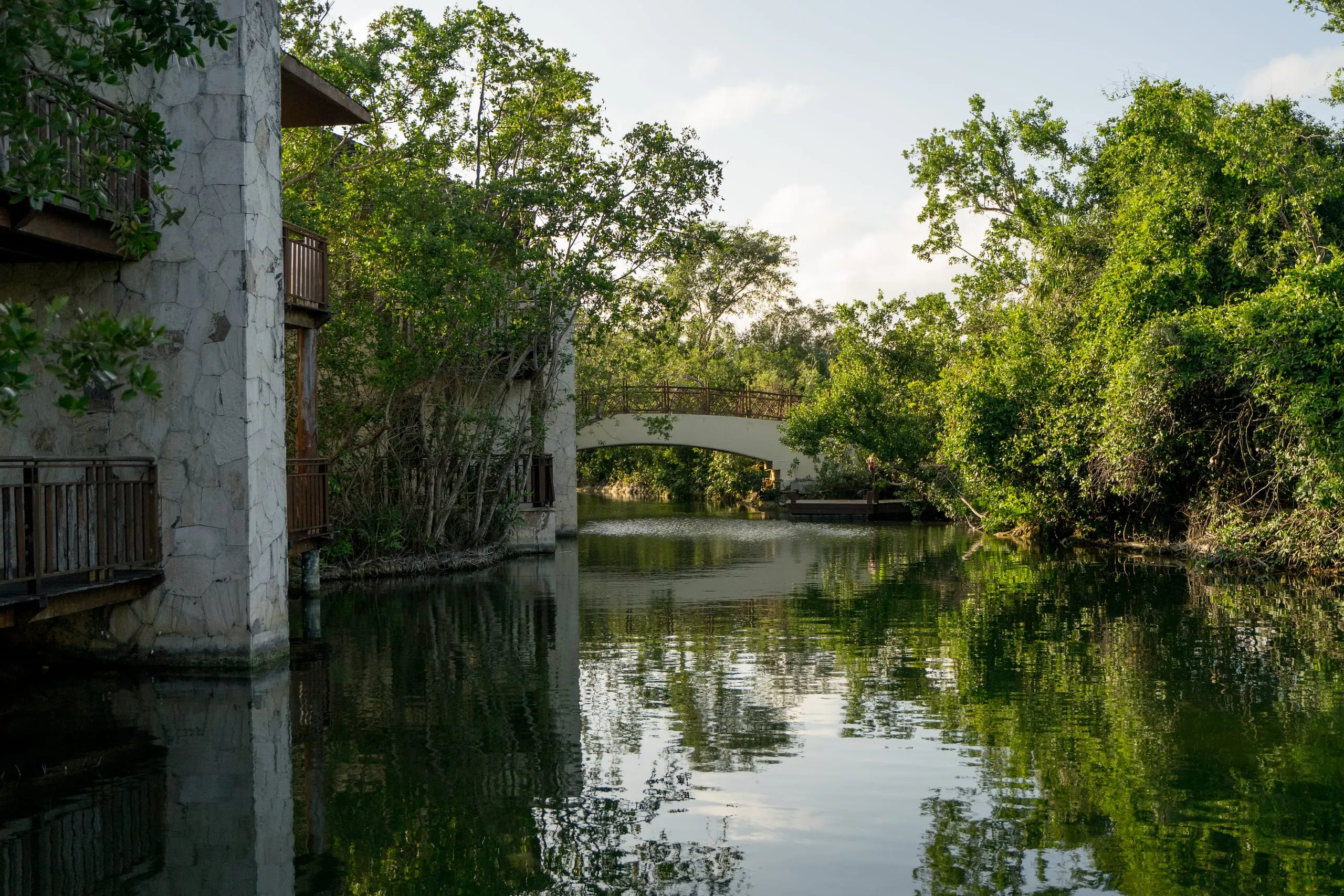 An arched footbridge crosses a still green canal framed by stone buildings and dense trees in Playa del Carmen.