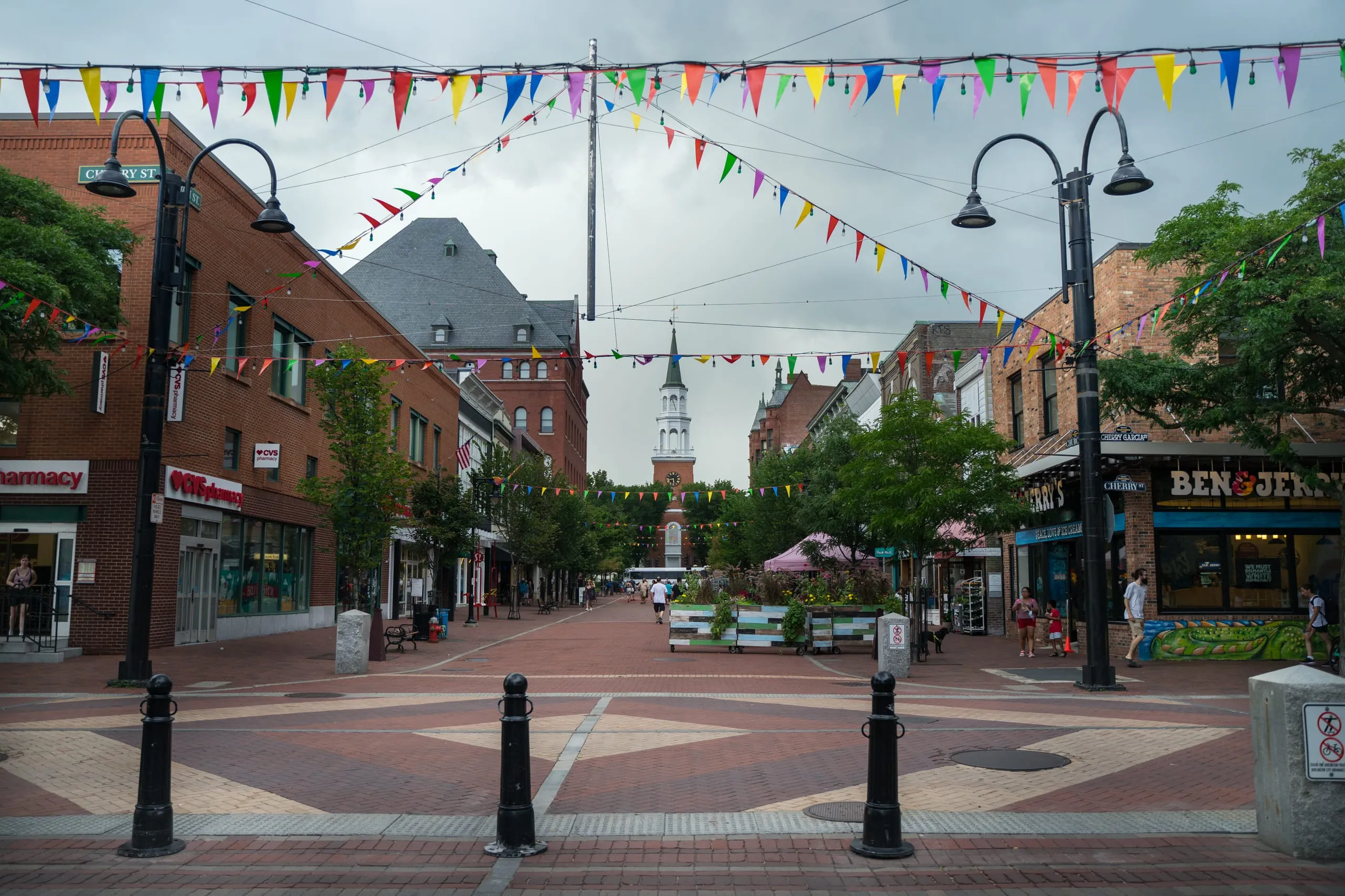 A brick pedestrian marketplace in Vermont strung with colorful pennants, shops on both sides and a white church steeple down the block.