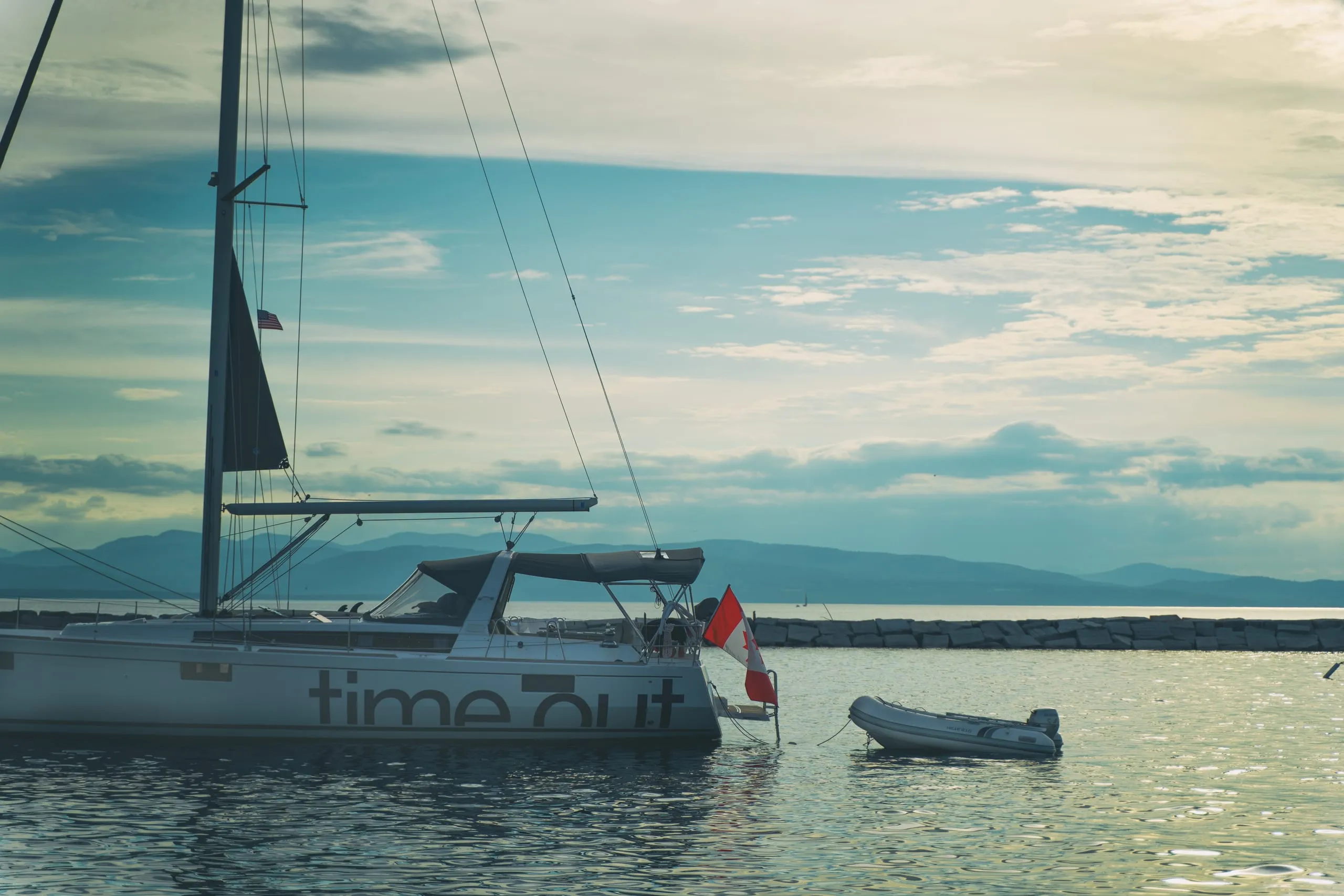 A moored sailboat named Time Out with a dinghy on a calm Vermont lake, mountains beyond.