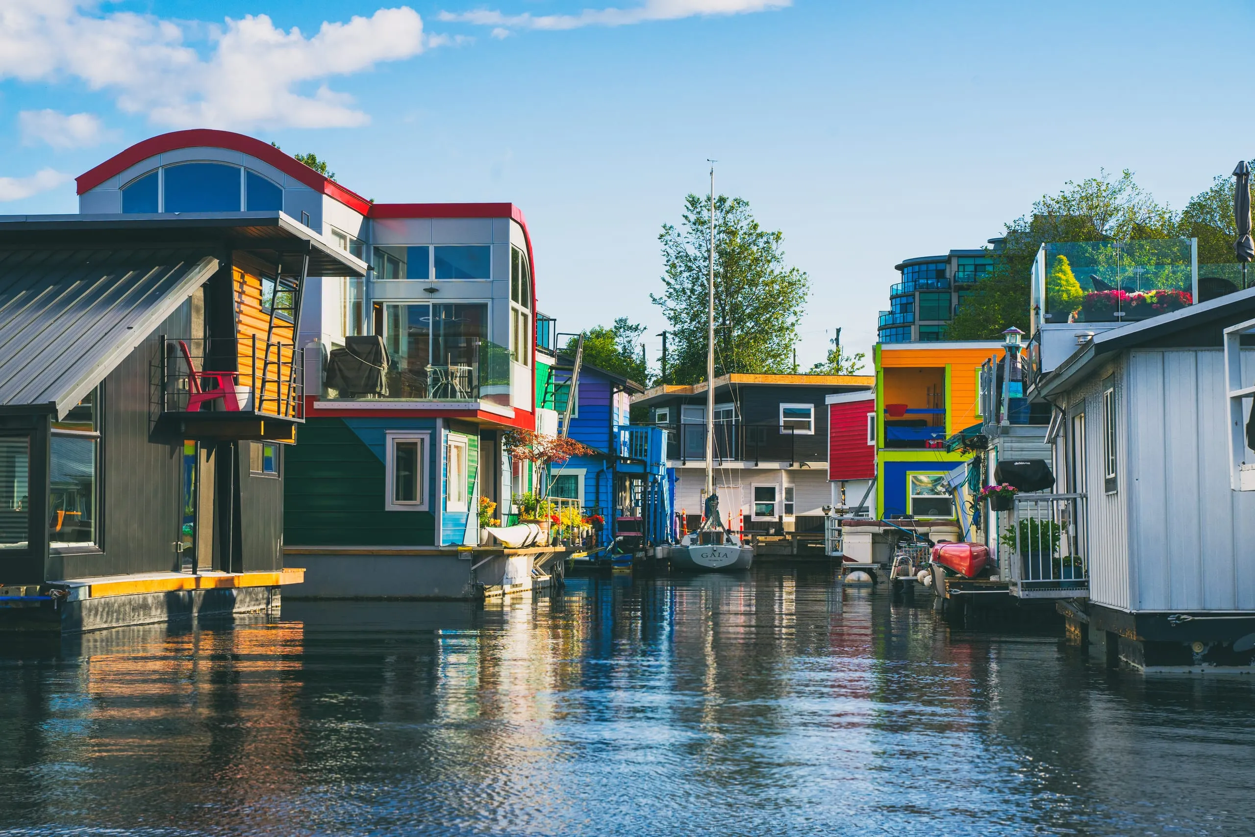 Brightly painted floating houses line a calm marina channel with a moored sailboat under blue sky in Victoria.