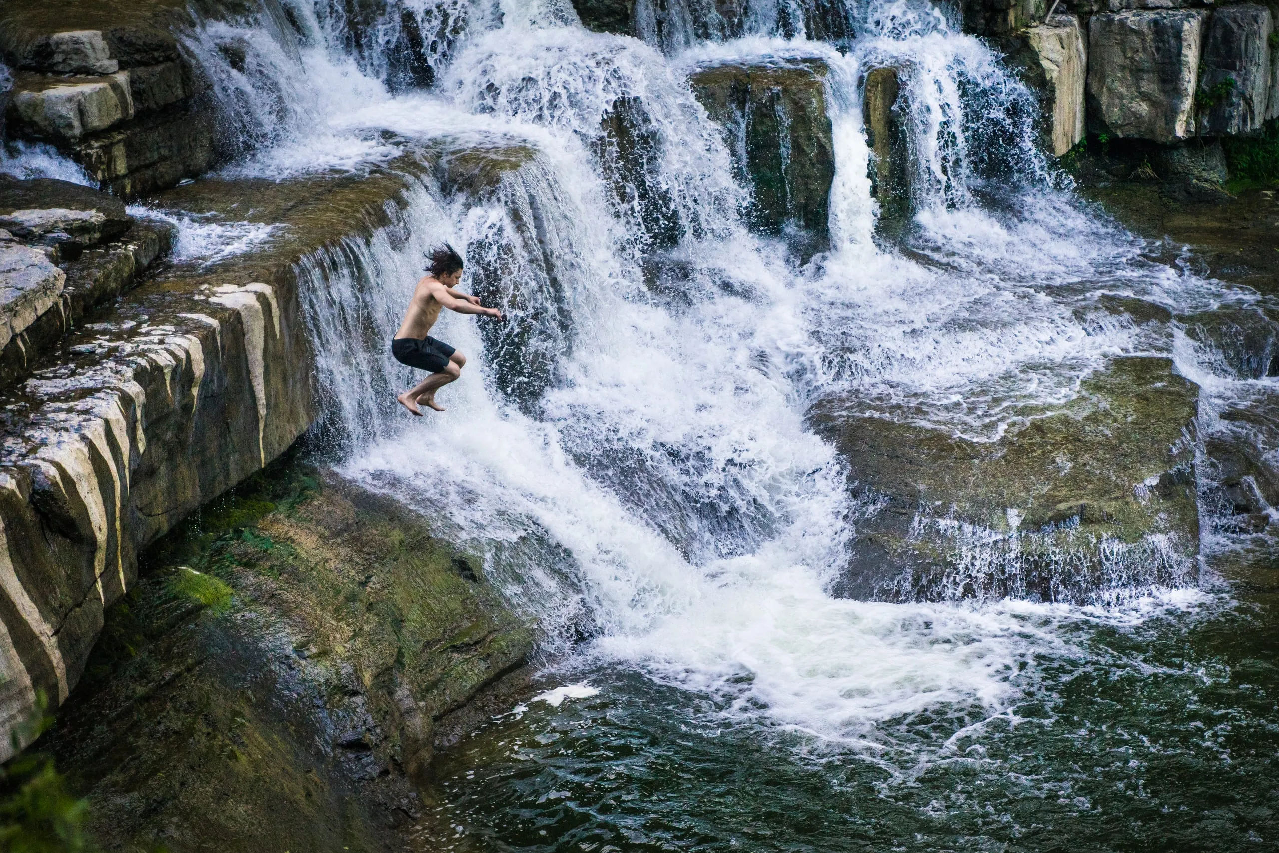 A shirtless man in black shorts jumps off a rock ledge toward the pool beside a cascading waterfall in the Finger Lakes.