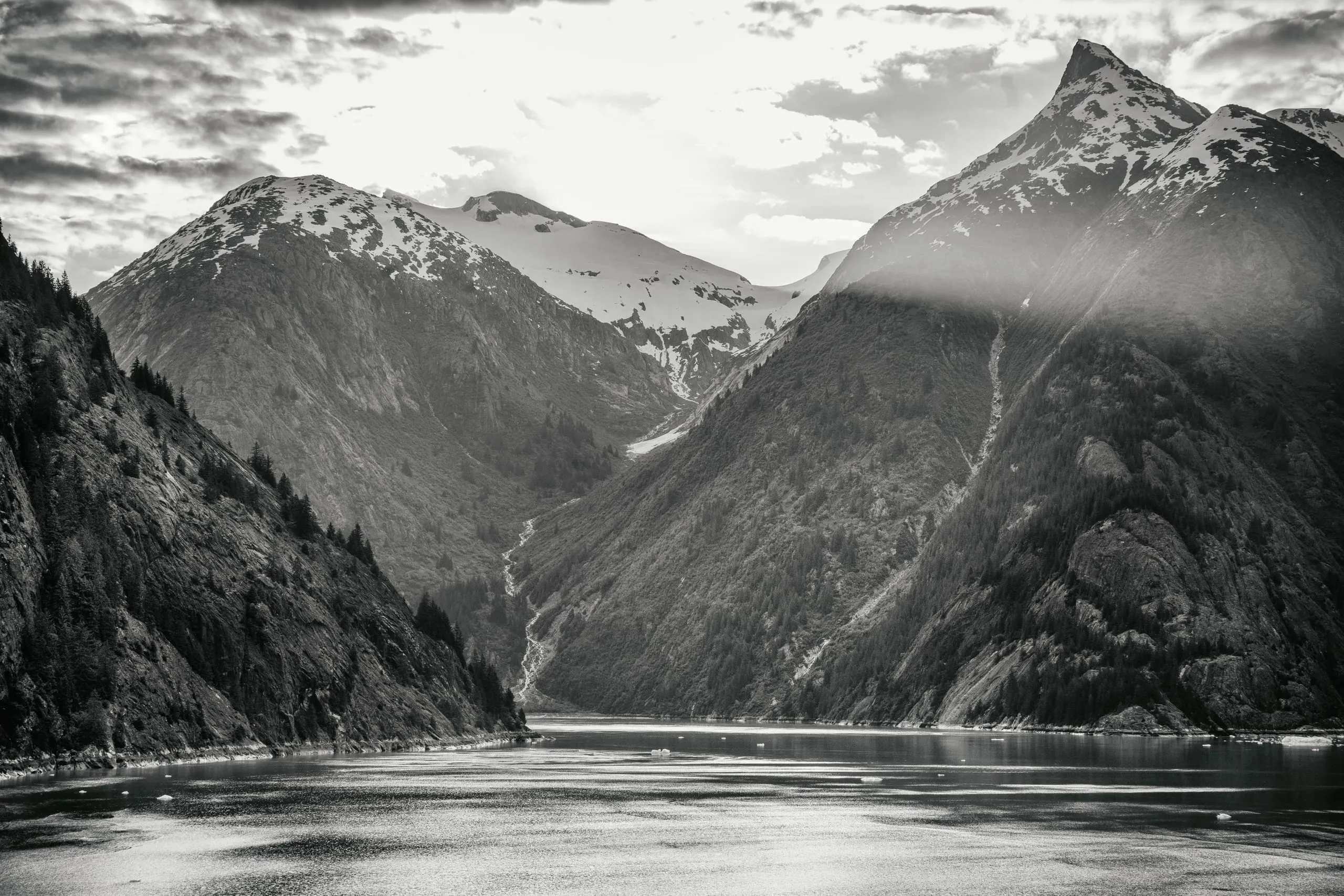 Black-and-white view down a narrow fjord flanked by steep peaks, with a backlit snow-patched summit and scattered ice on dark water.