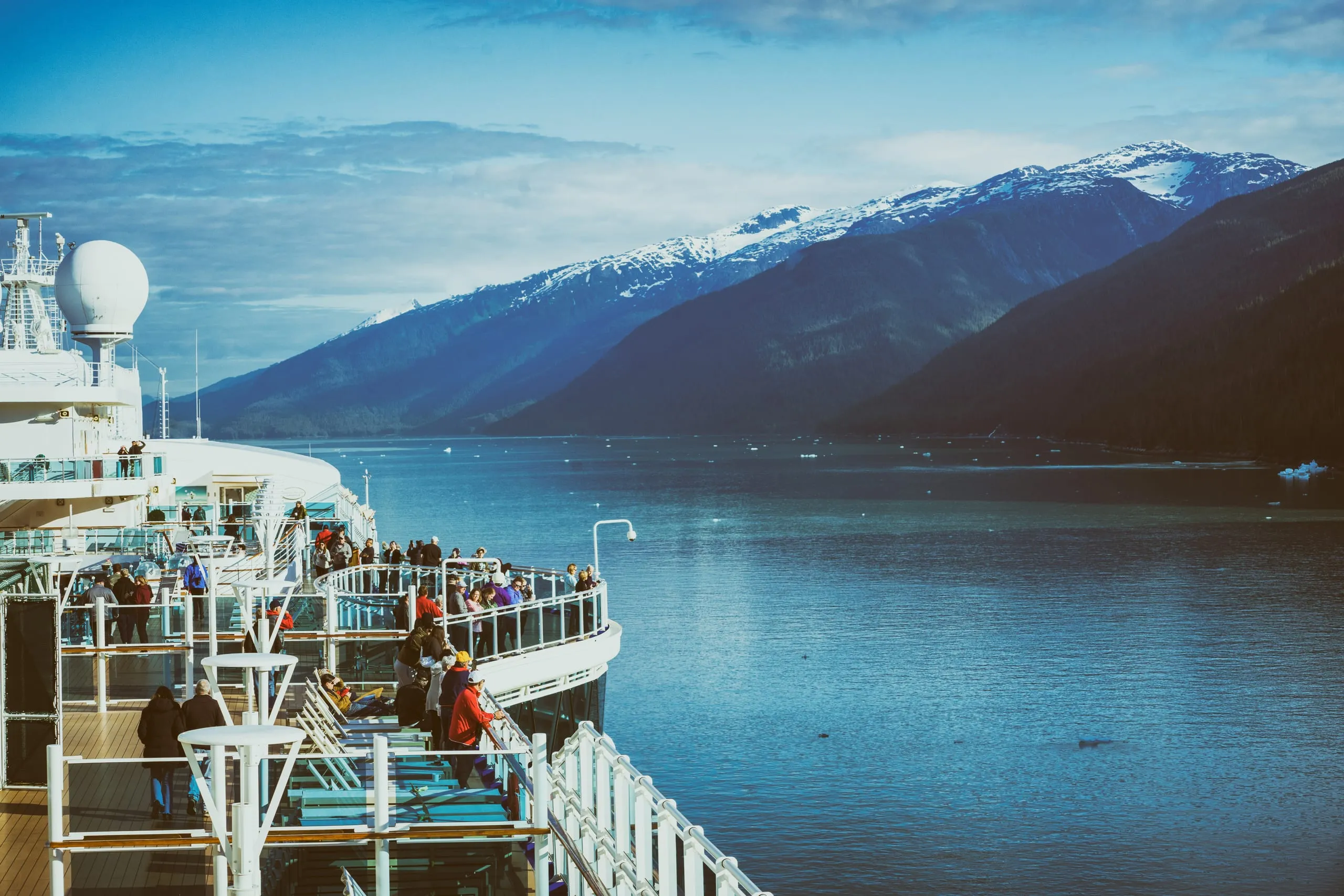 Passengers line the curving railings and lounge chairs on a cruise ship's upper deck as it sails a calm fjord toward snow-capped mountains.
