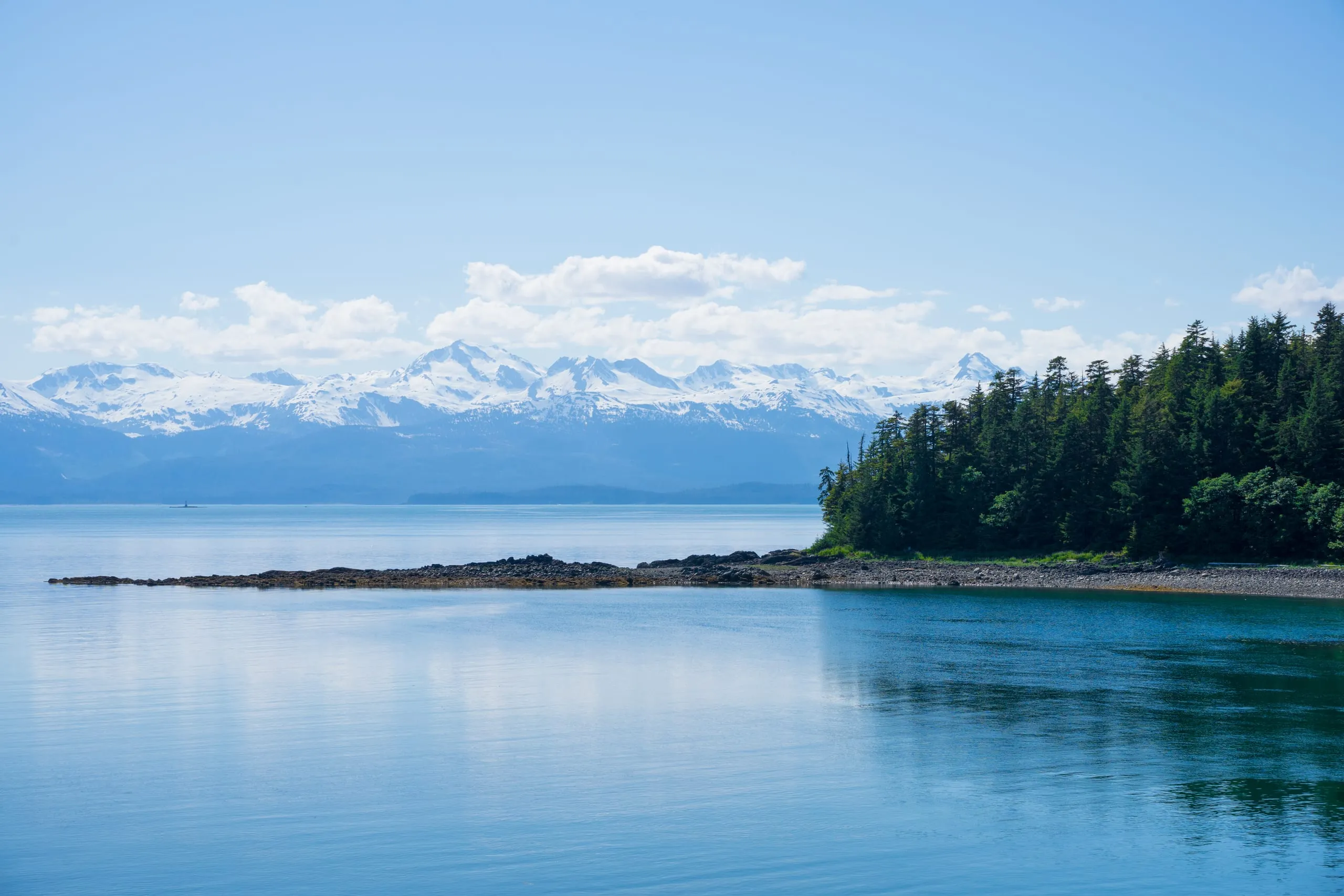 A still turquoise inlet with a rocky spruce-covered point on the right and a long snow-capped mountain range across the horizon.