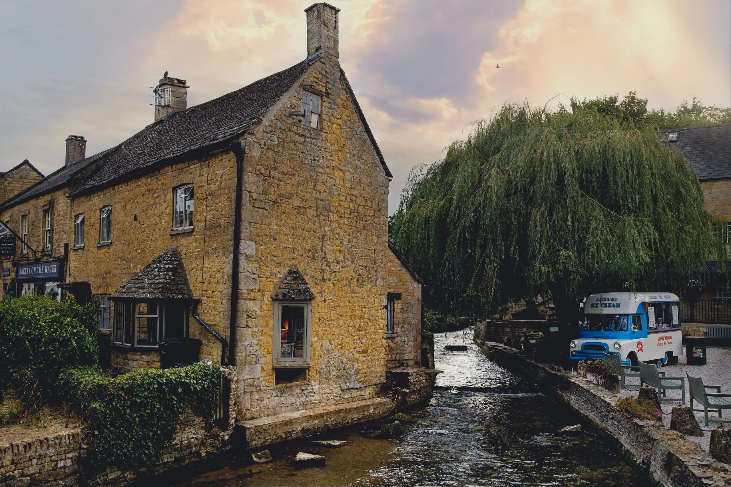 A stone cottage marked 'Bakery on the Water' beside a narrow stream, with a weeping willow and a vintage ice cream van on the opposite bank at dusk, in Bourton-on-the-Water, England.