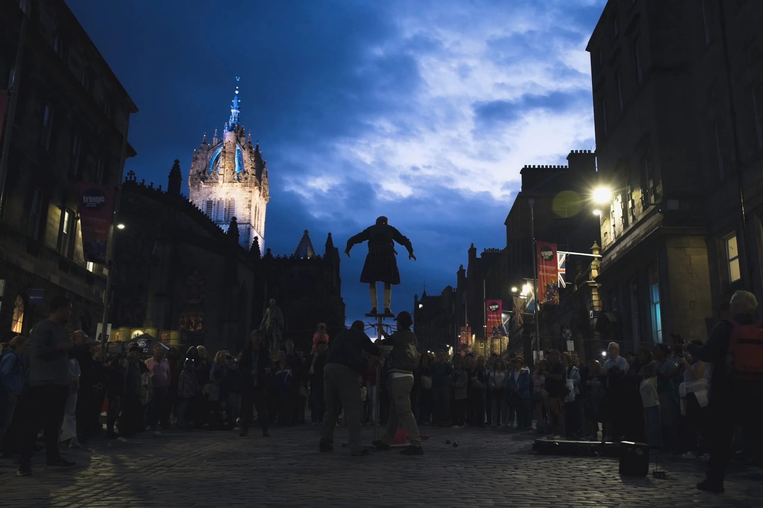 A color photograph of a kilted street performer balanced atop a narrow pole on Edinburgh's Royal Mile at dusk, silhouetted against the illuminated crown spire of St Giles' Cathedral as a crowd watches below.
