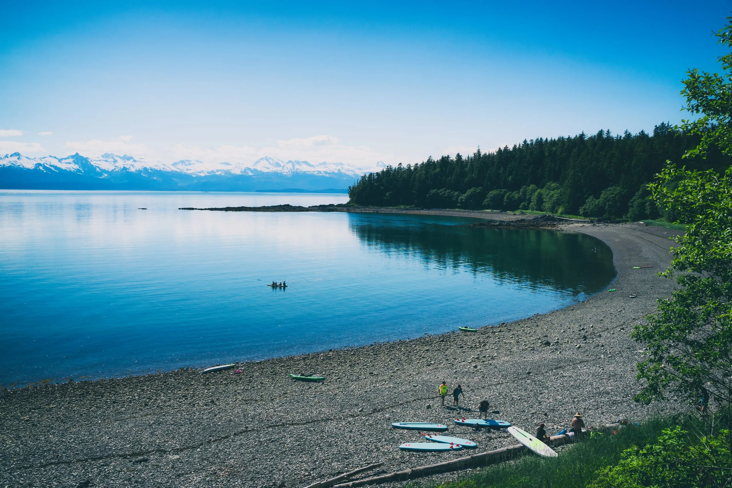 Kayaks and paddleboards lie on a gray pebble beach along a calm cove where paddlers float, with forested shore and distant snowy peaks.