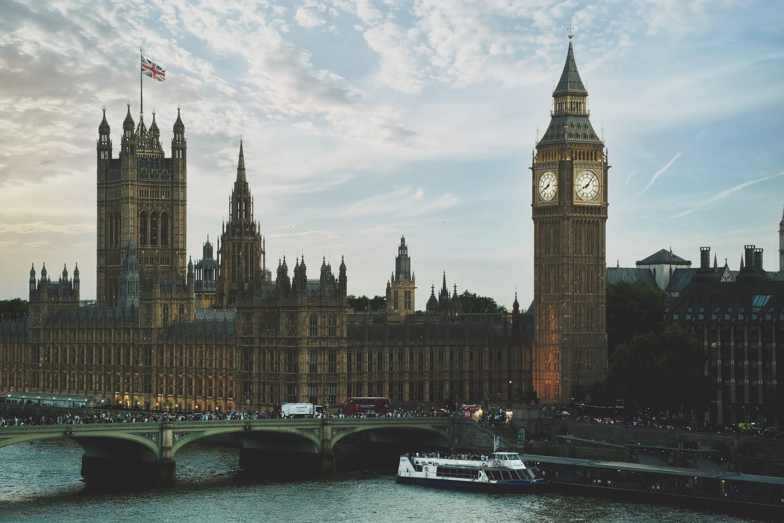 Photograph of Big Ben's illuminated clock tower and the Houses of Parliament above the River Thames at dusk, with Westminster Bridge and a river cruise boat in the foreground, London, England.