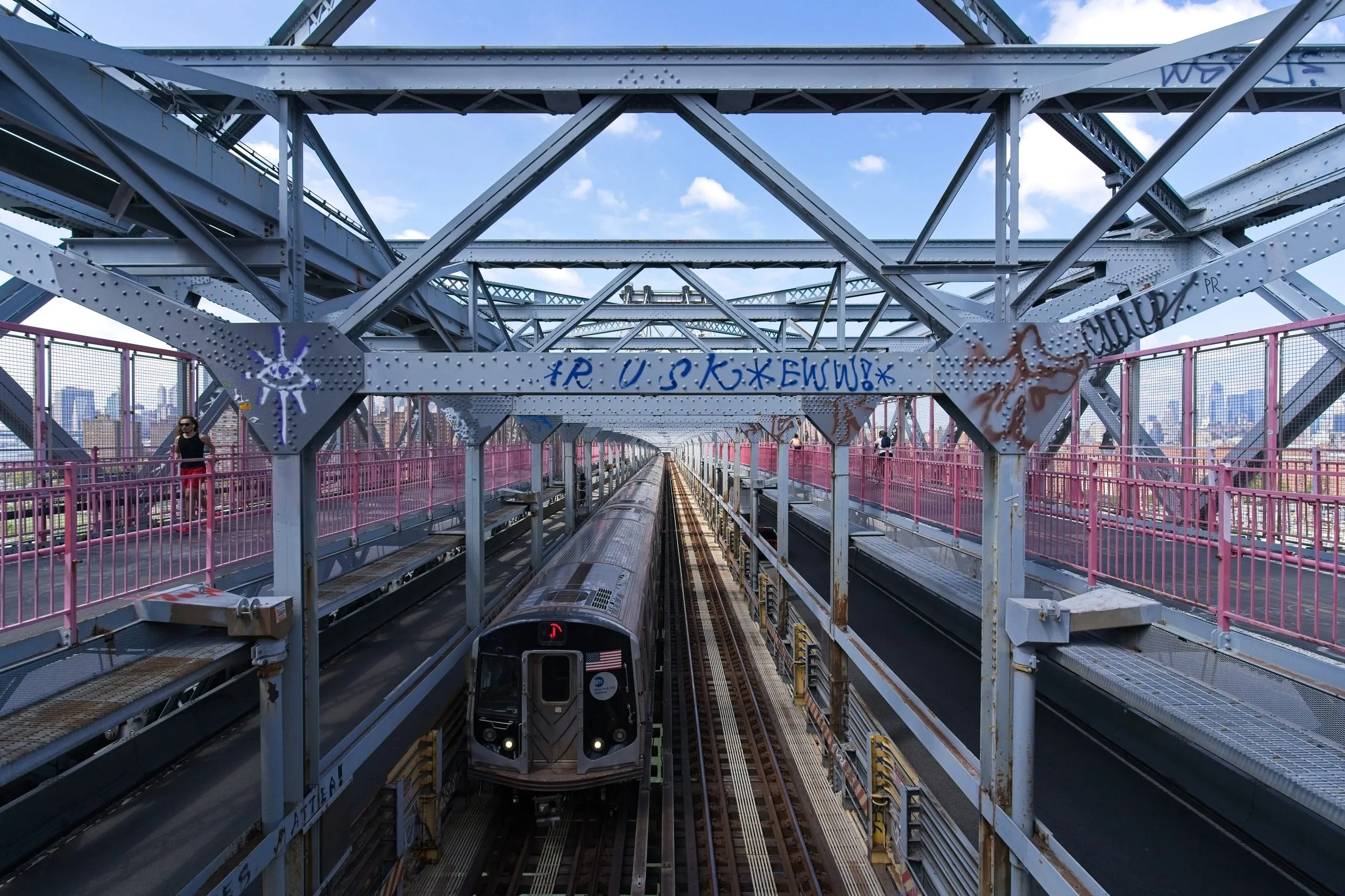 A color photograph of a New York City subway train crossing the Williamsburg Bridge on elevated tracks, framed by steel girders and pink pedestrian railings with the Manhattan skyline visible beyond.