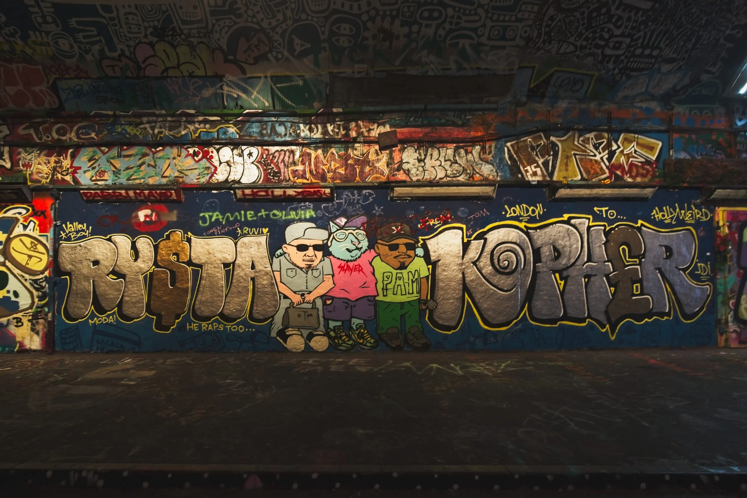 Photograph of a large graffiti mural in London reading 'RYSTA KOPHER' in bold bubble letters, with three cartoon figures in sunglasses standing between the words.