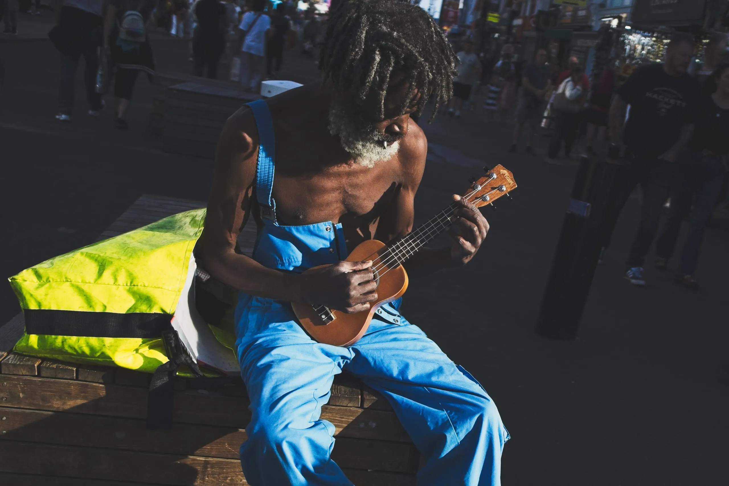 A close-up photograph of a street musician in blue overalls bent over a ukulele, seated on a bench in Camden, London, England.