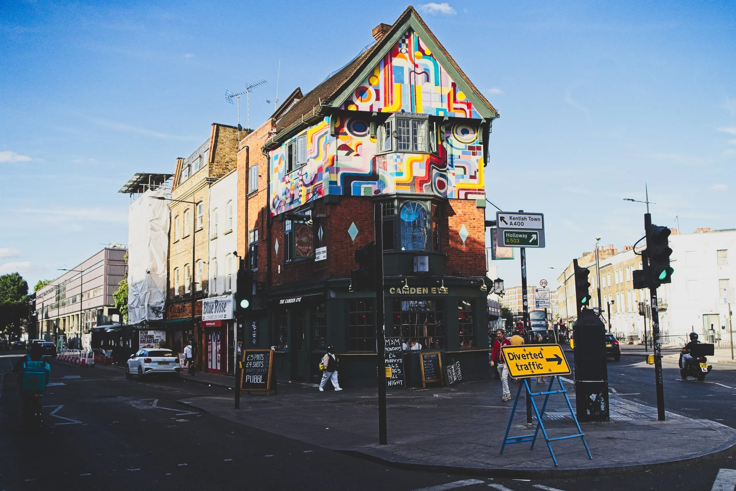 A photograph of a triangular corner pub named The Camden Eye, its upper stories covered in a bold geometric mural, at a street corner in Camden, London, England.