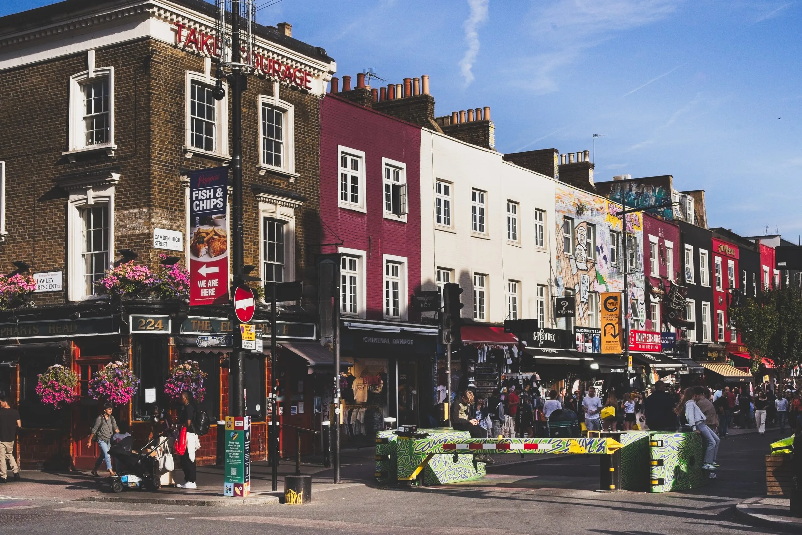 A photograph of colorful row houses and shopfronts along Camden High Street in London, England, with hanging flower baskets, pedestrians, and a graffiti-painted traffic barrier in the foreground.