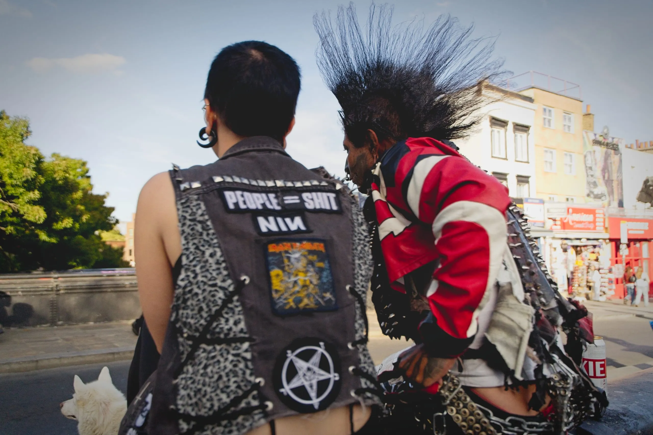 Color photograph of two punks seen from behind on a street in Camden, London, one in a studded vest covered in band patches, the other with a tall black mohawk and spiked jacket.