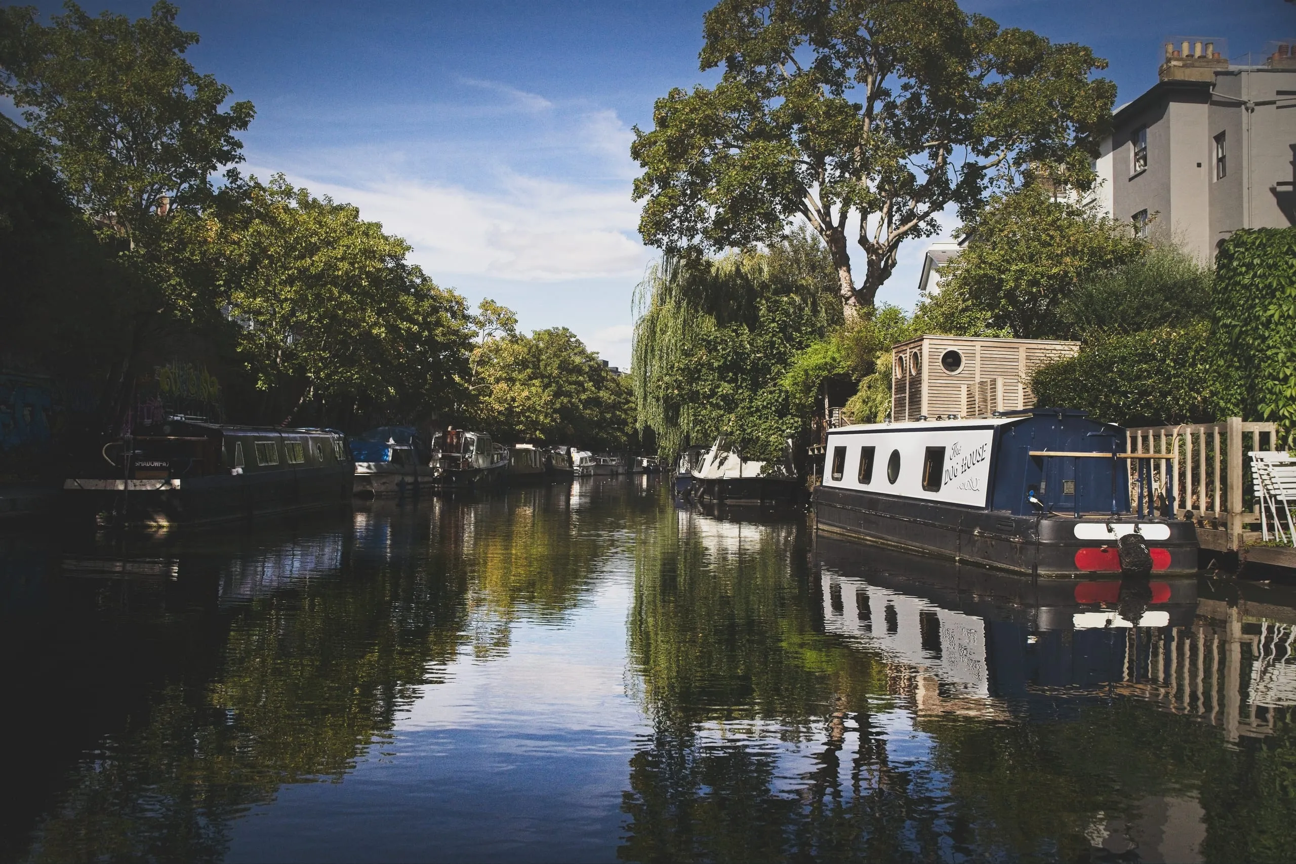 Color photograph of a tree-shaded stretch of Regent's Canal in London, blue sky reflected in the still water, with houseboats moored along the bank.