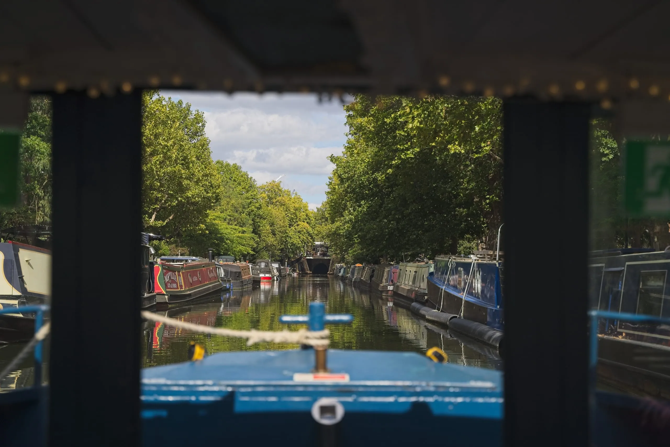 Color photograph of a tree-canopied canal in London lined with moored narrowboats, framed by the dark structure of the boat it was taken from, a tunnel visible ahead.