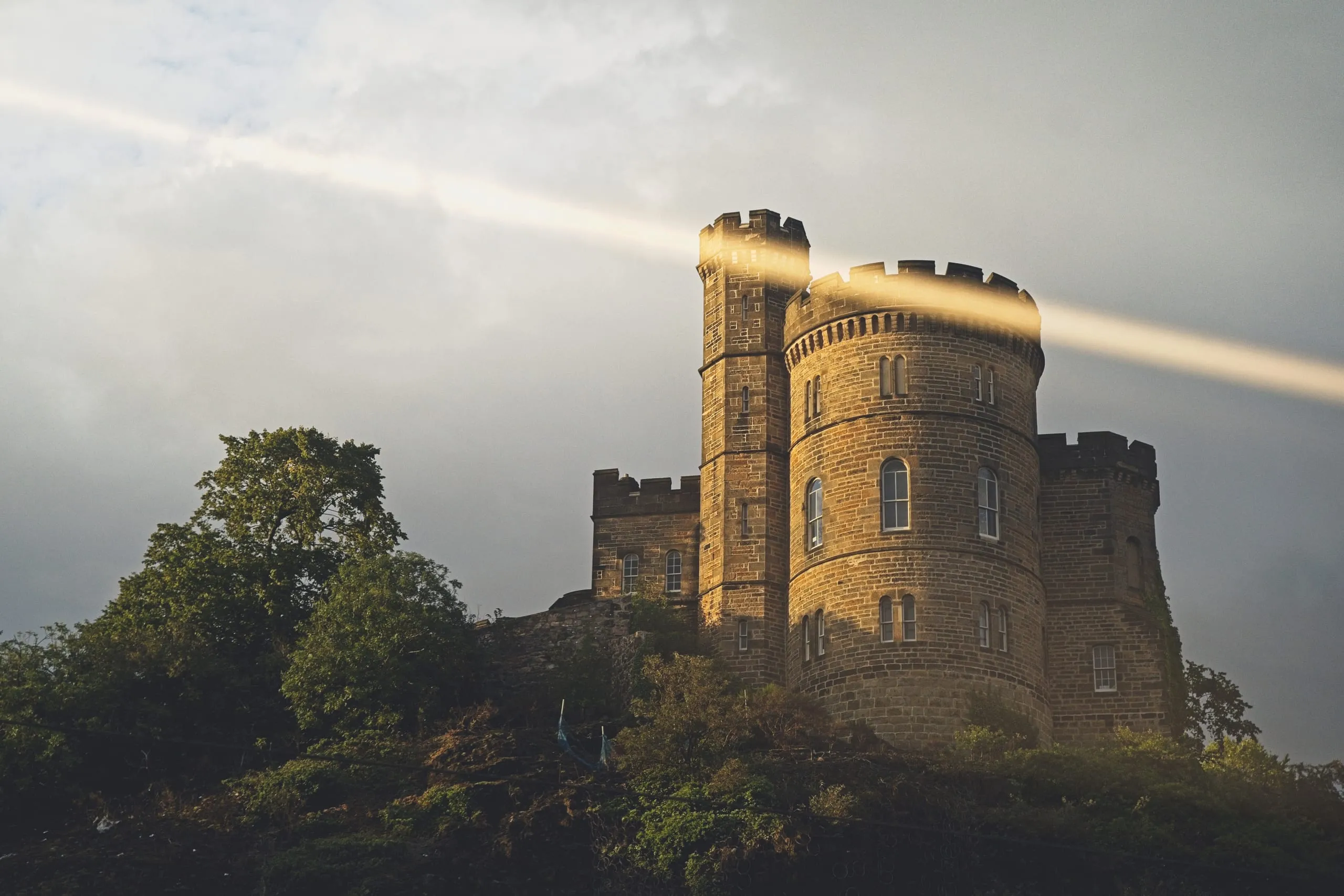 A color photograph of a crenellated castellated stone building, resembling Edinburgh's Calton Hill architecture, lit by a dramatic shaft of sunlight through cloudy skies.