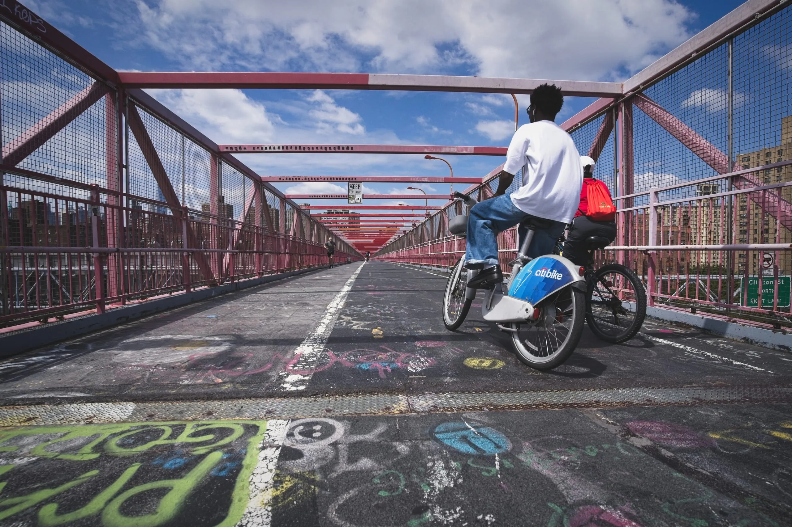 A color photograph of two cyclists riding Citi Bikes down the graffiti-covered pedestrian path of the Williamsburg Bridge, with red pipe railings and the Brooklyn skyline in the distance.