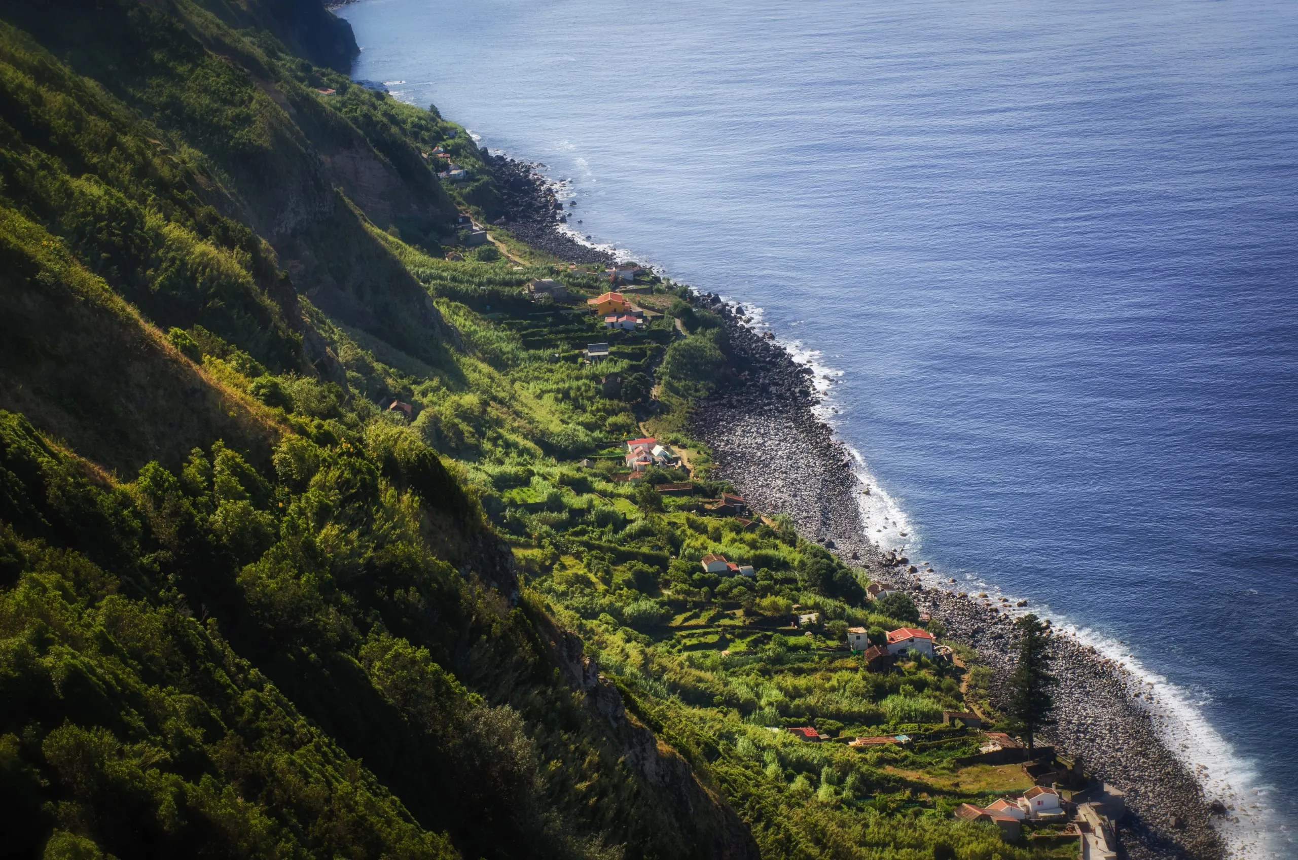 Aerial view down a steep terraced green cliffside dotted with scattered farmhouses meeting a long boulder-strewn shoreline and deep blue sea.