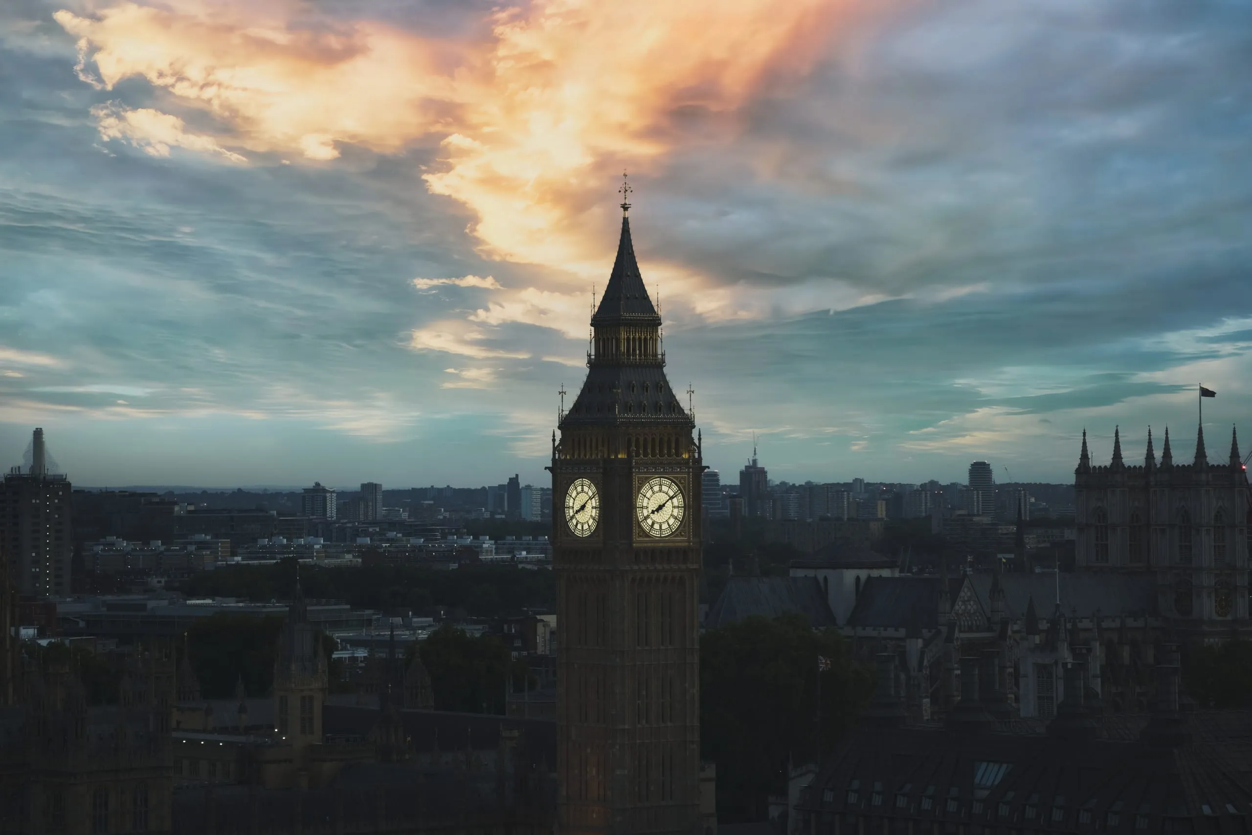 Photograph of Big Ben's illuminated clock face rising above the London skyline at dusk, beneath a sky streaked orange and blue, London, England.