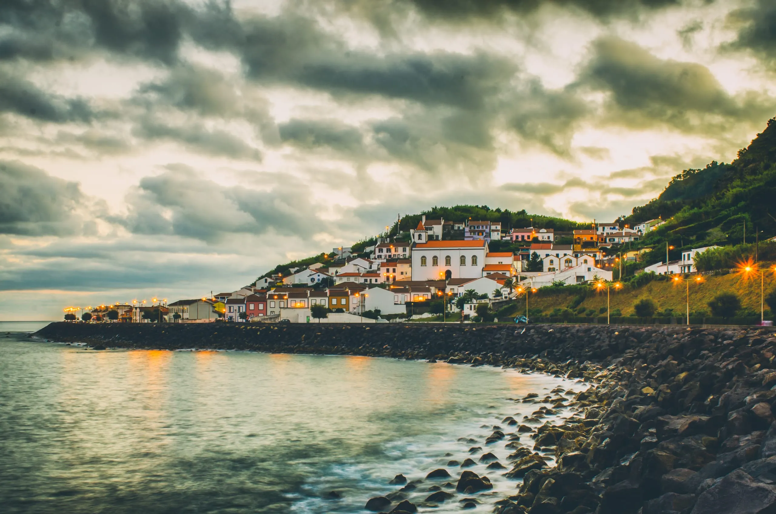 A hillside coastal village of white houses and a church glows with street and window lights at dusk above a rocky shoreline.