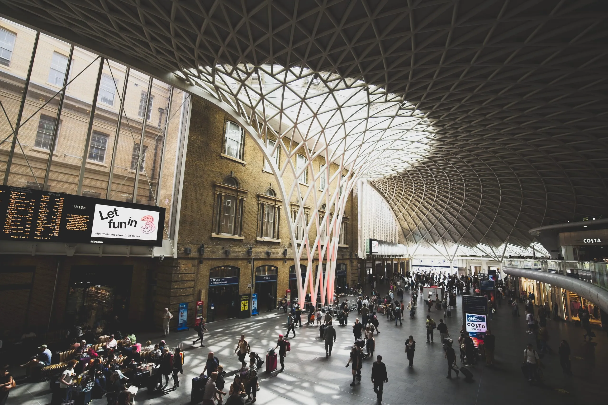 A photograph of the busy concourse inside King's Cross station in London, with the latticed glass roof overhead and travelers pulling luggage across the floor.