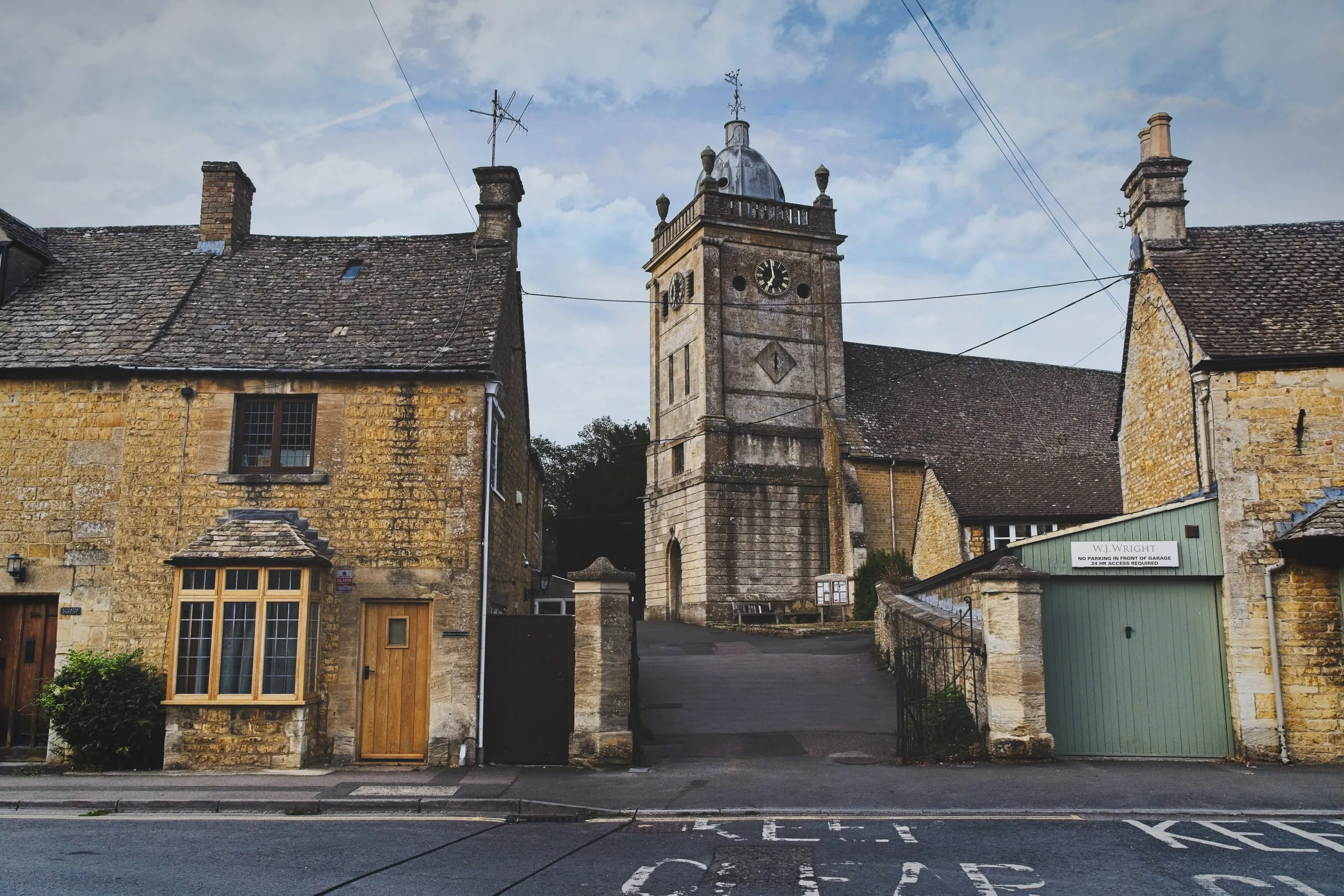 A stone clock tower with a domed cupola rises above a row of honey-colored cottages on a quiet Cotswolds village street, England.