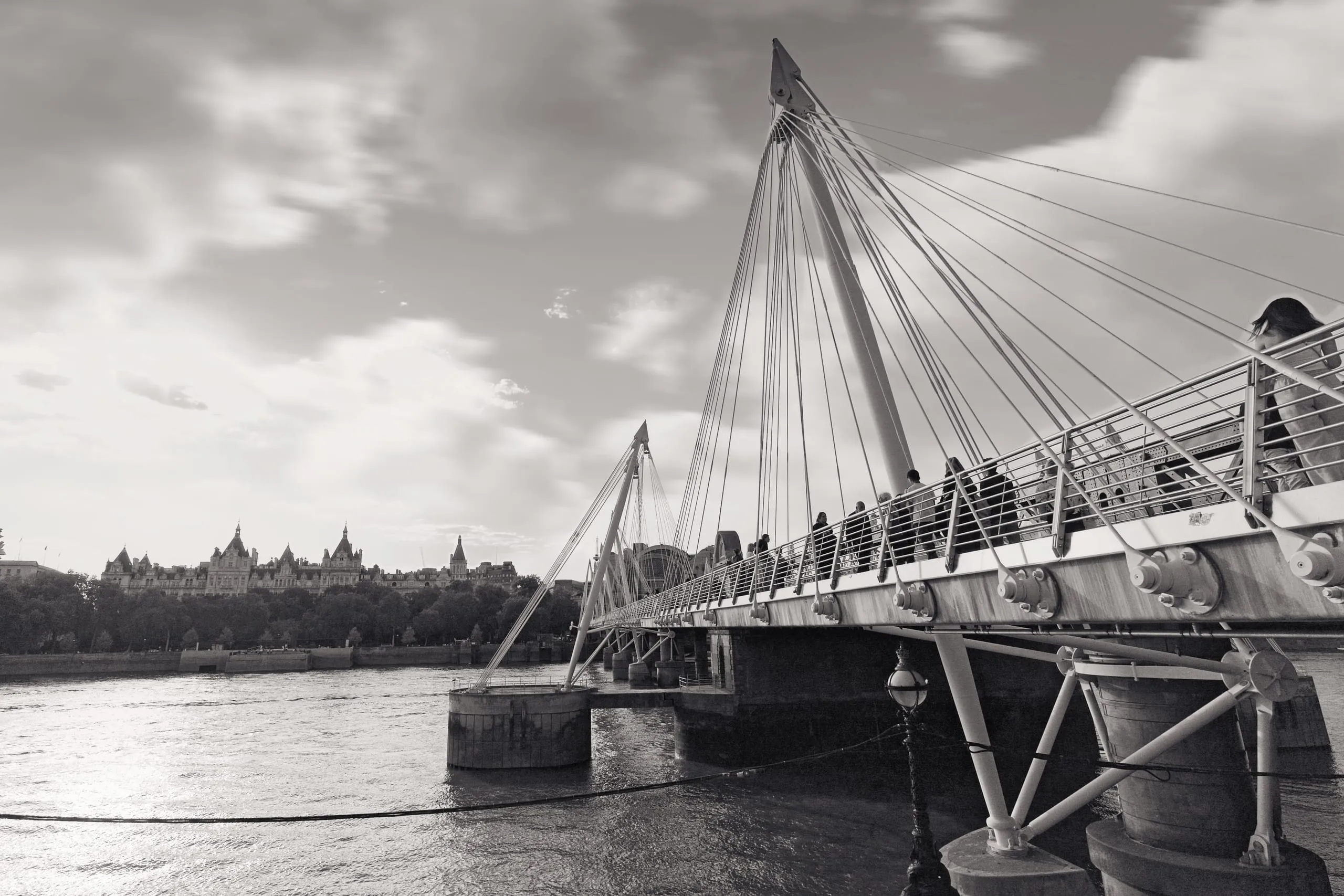 Black and white photograph of pedestrians walking across the Golden Jubilee footbridge over the River Thames, with the ornate rooftops of Whitehall Court visible on the far bank, London, England.