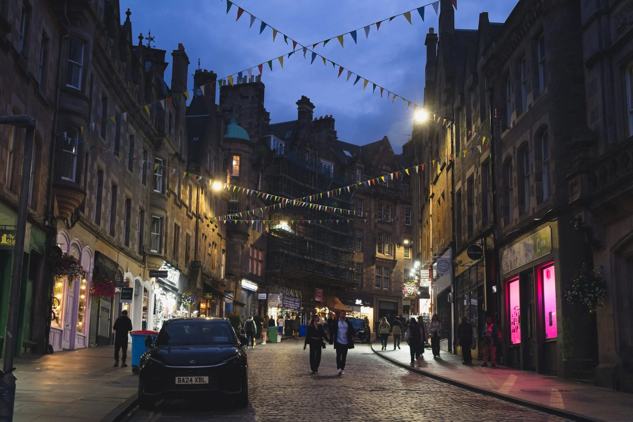 A color photograph of Victoria Street in Edinburgh's Old Town at dusk, its curved row of painted shopfronts strung with triangular bunting as pedestrians stroll the cobbles under a darkening sky.