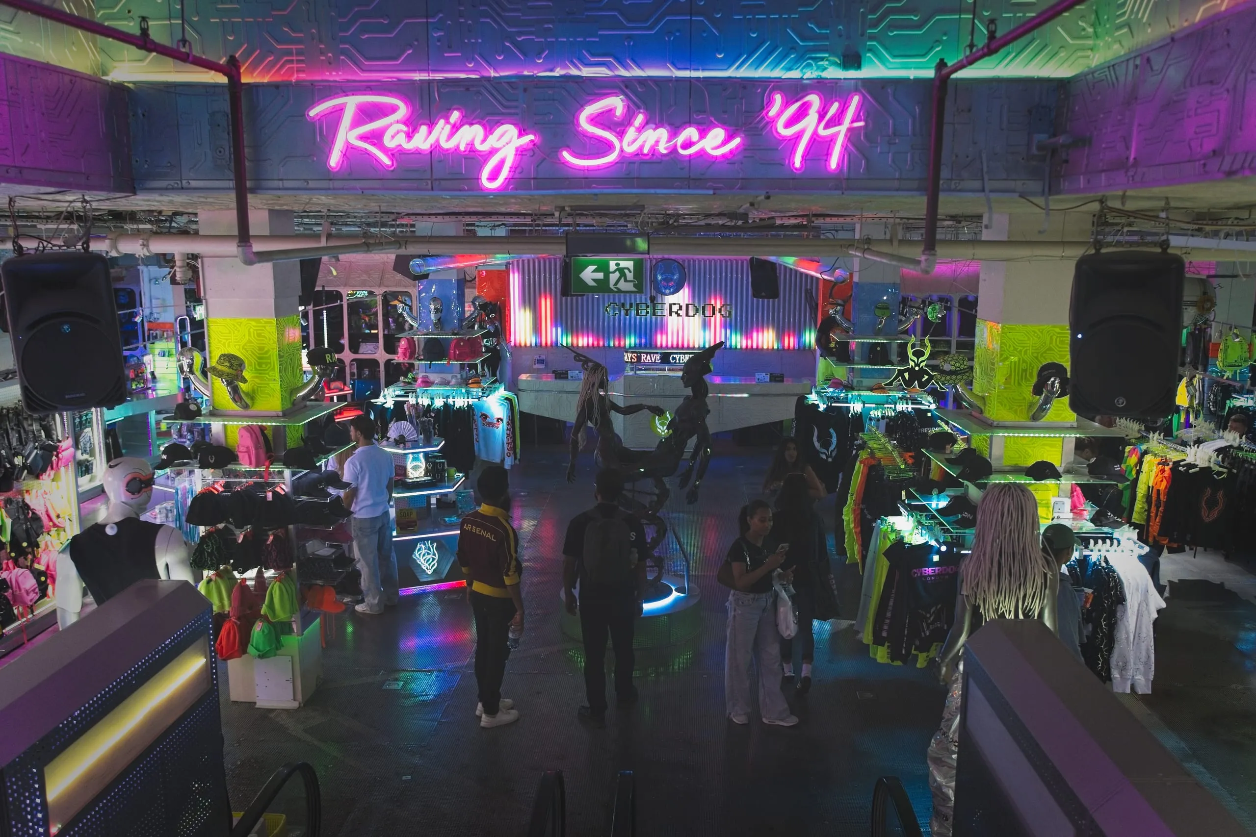 Color photograph of the neon-lit interior of the Cyberdog store in Camden Market, London, with a pink 'Raving Since '94' sign glowing above shoppers and clothing racks.