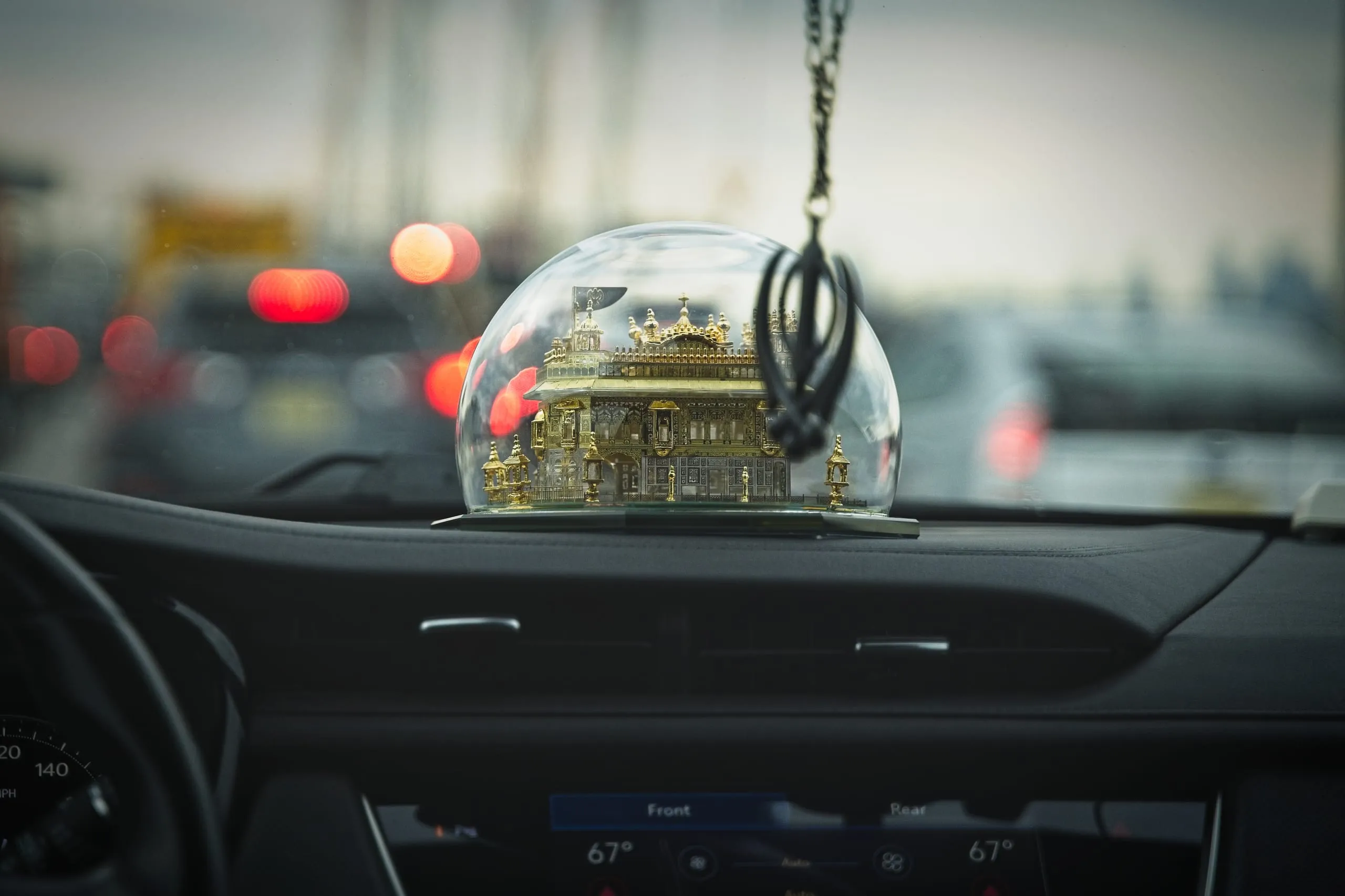 A color photograph of a small golden temple ornament under a glass dome on a car dashboard, with blurred red taillights of Brooklyn traffic visible through the windshield at dusk.