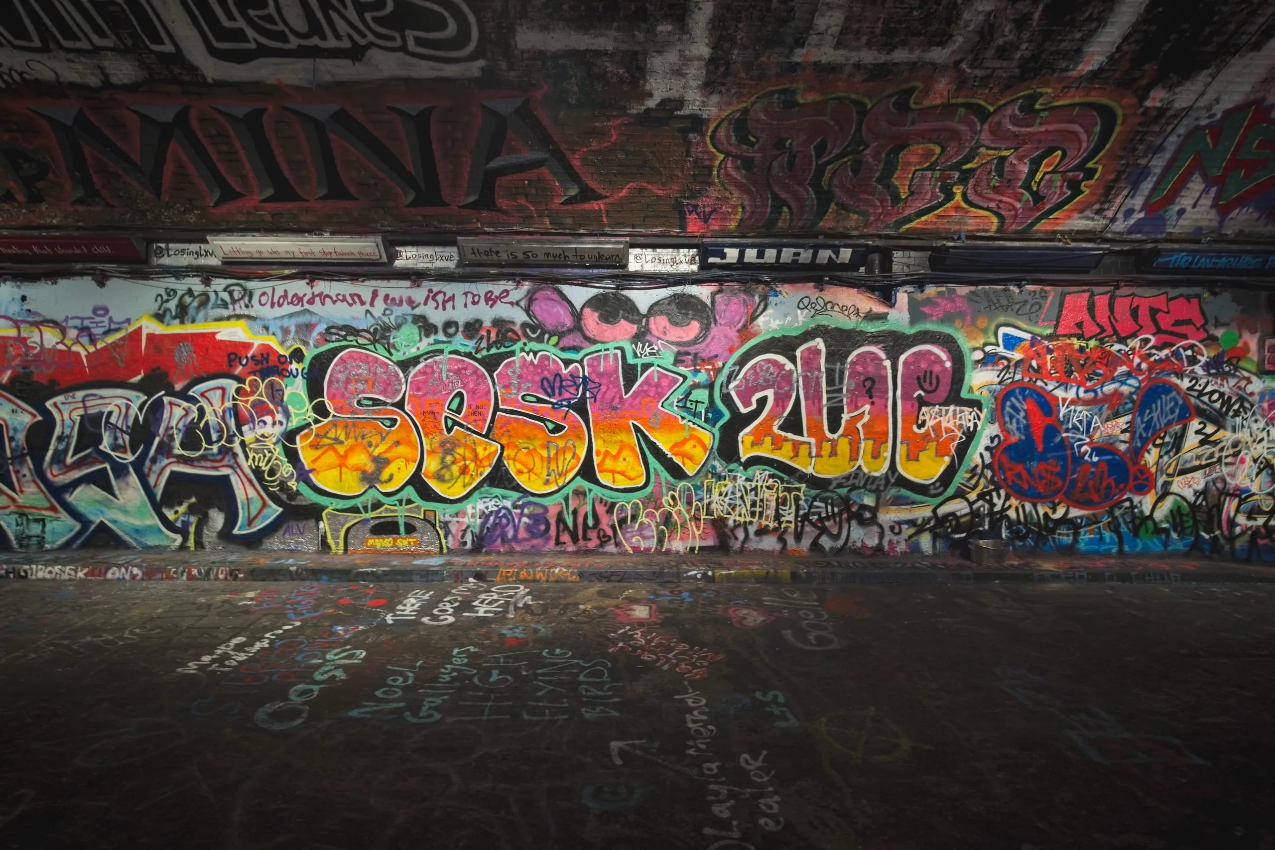 Photograph of a densely tagged graffiti wall in London covered in overlapping spray-painted names and slogans, with handwritten messages chalked onto the pavement below including references to Oasis.