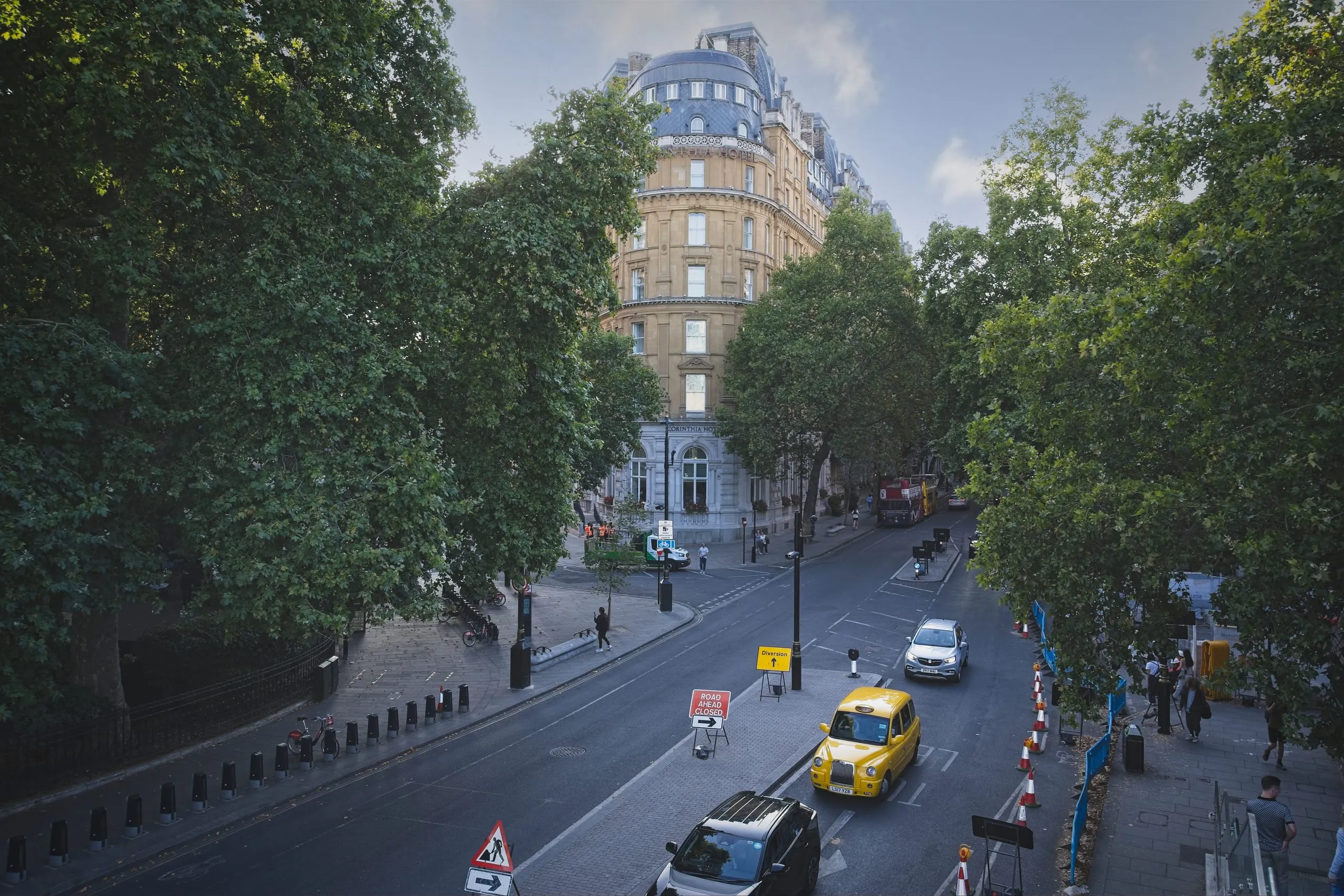 Photograph of a tree-lined London street leading toward the Corinthia Hotel, with a black taxi cab and road diversion signs in the foreground, London, England.