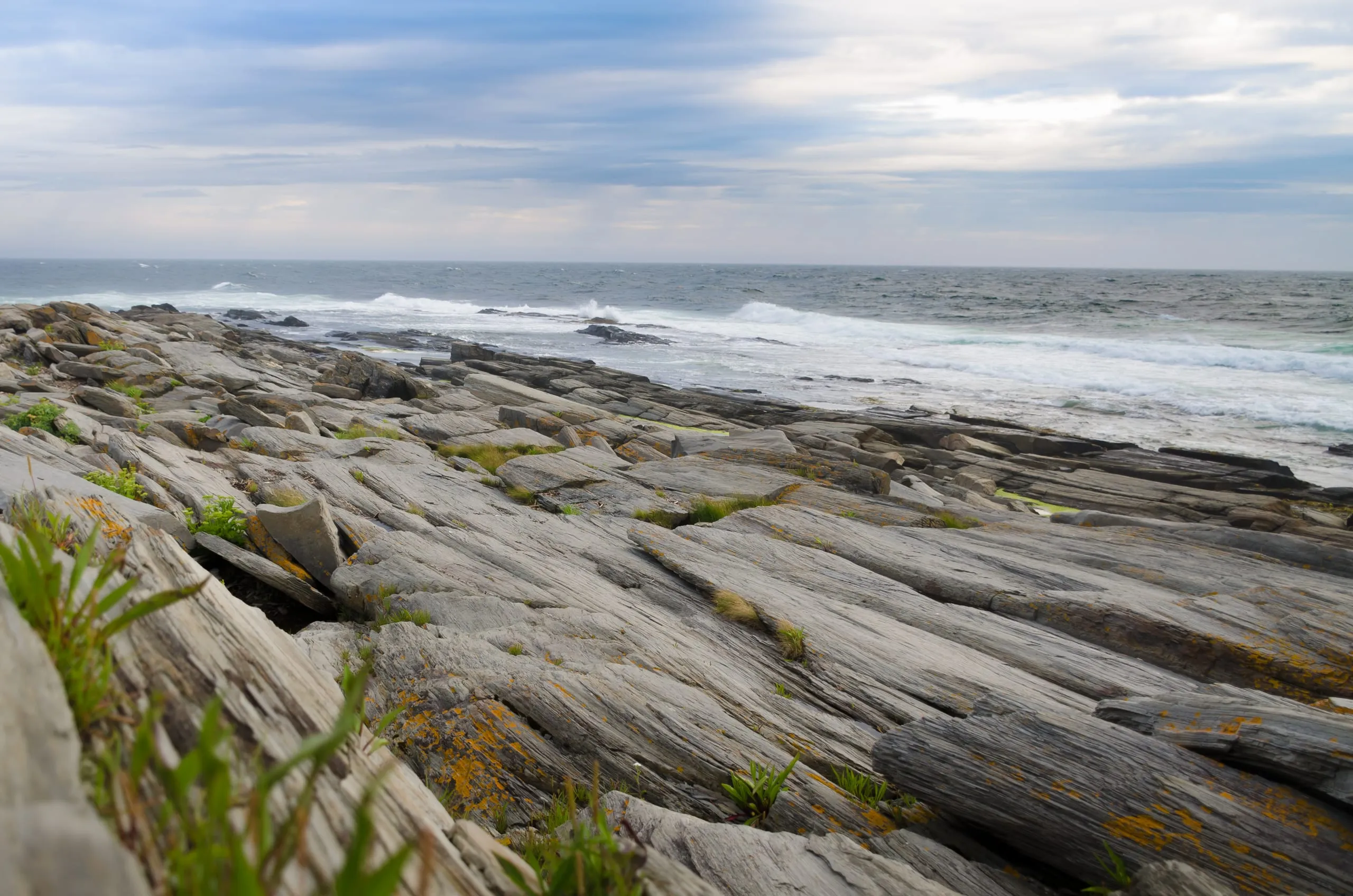 Striated gray rock ledges tilt down toward breaking surf under a cloudy sky on the Portland, Maine coast.