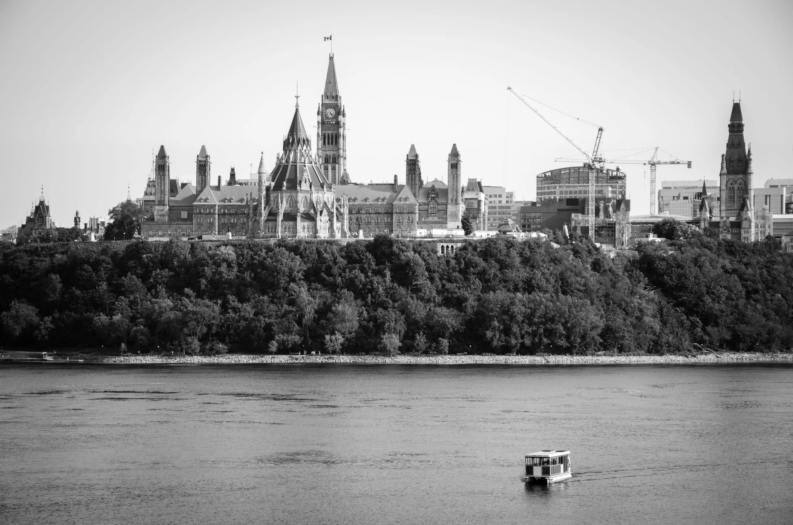 Black-and-white view of Ottawa's Parliament Hill and Peace Tower across the river as a small ferry glides past.