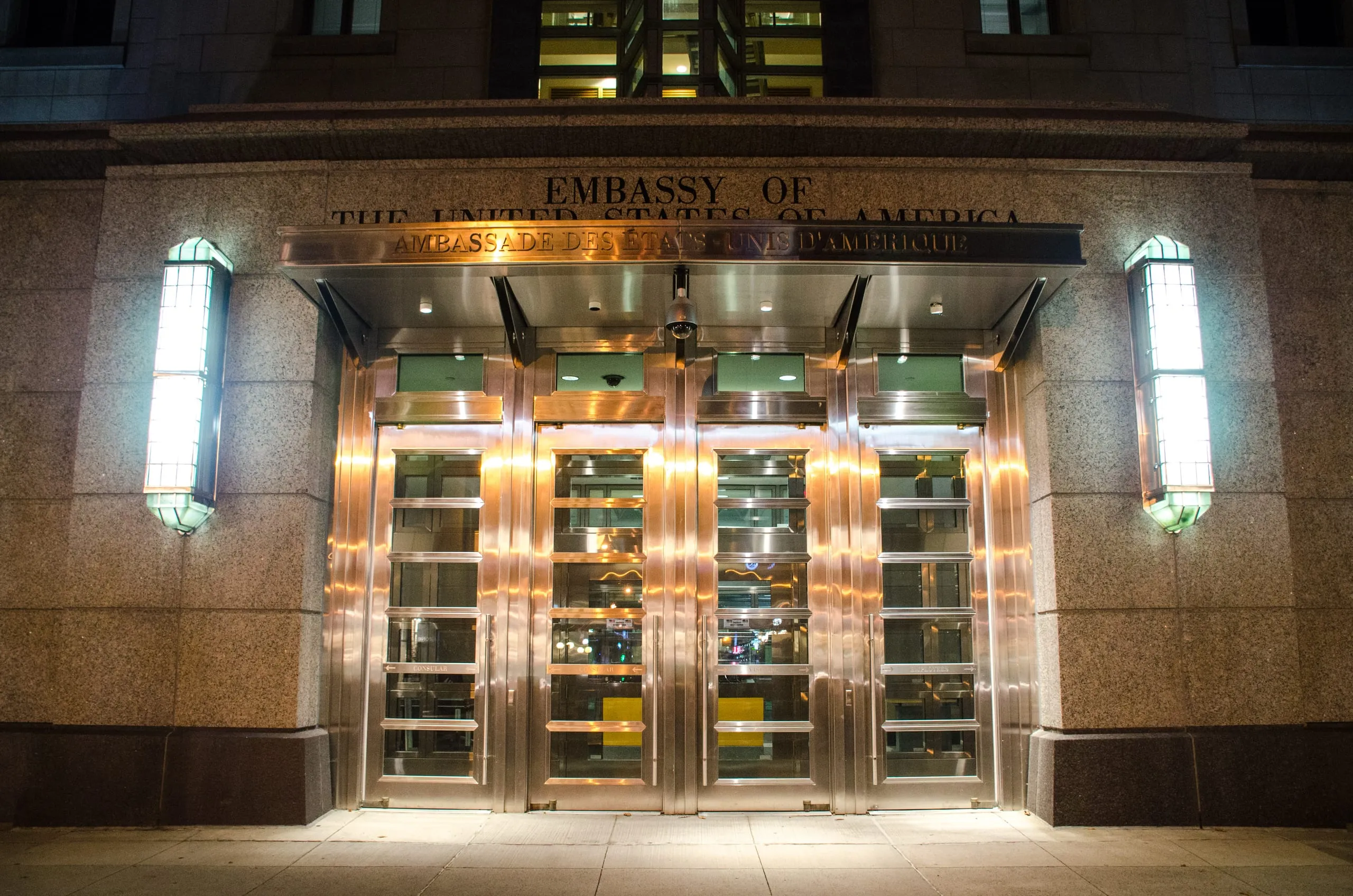 The lit brass entrance of the United States Embassy at night, with bilingual English and French lettering, in Ottawa.
