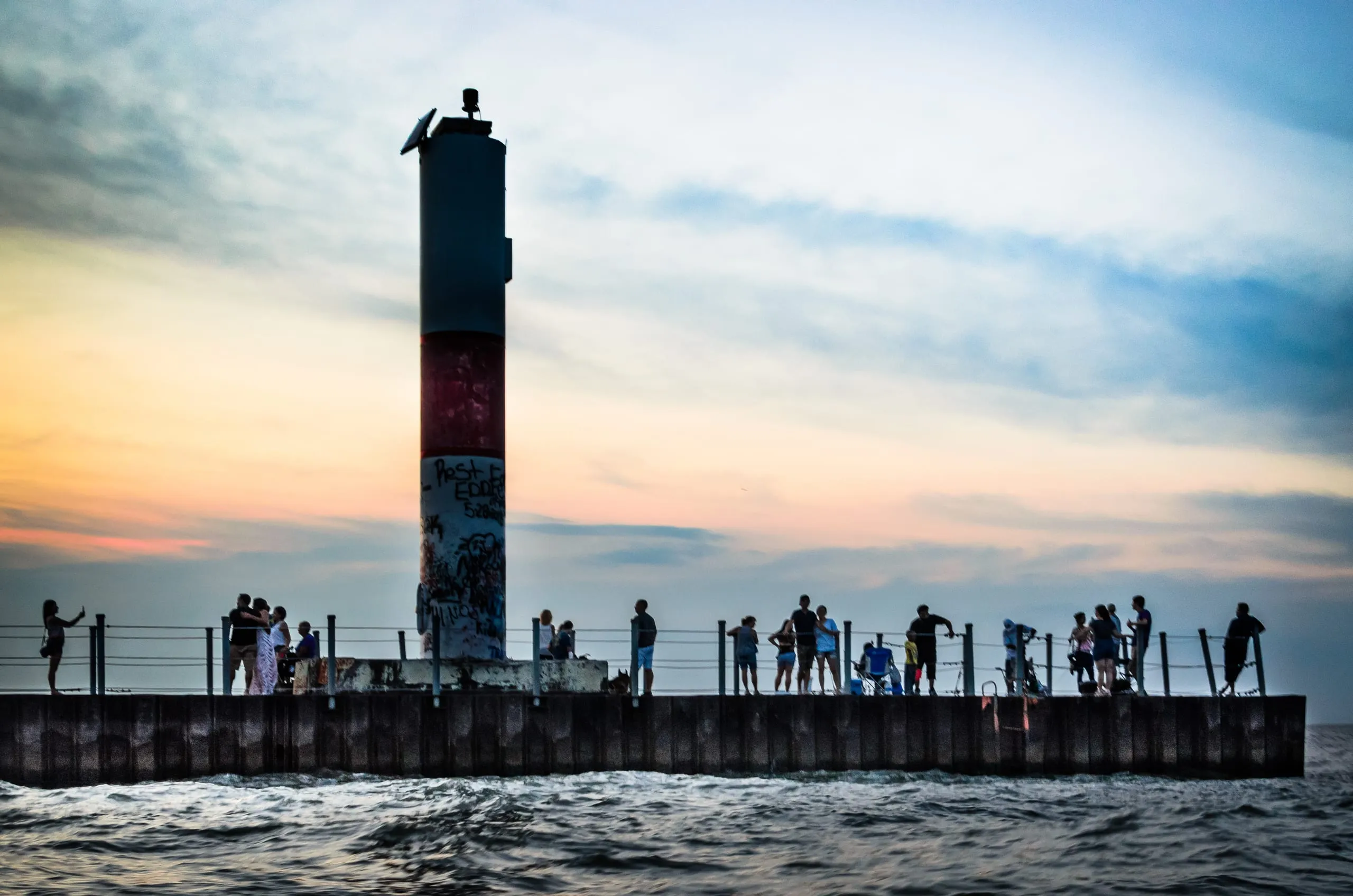 Silhouetted crowd lines a long pier beside a tall graffitied channel beacon at sunset in Rochester, NY.