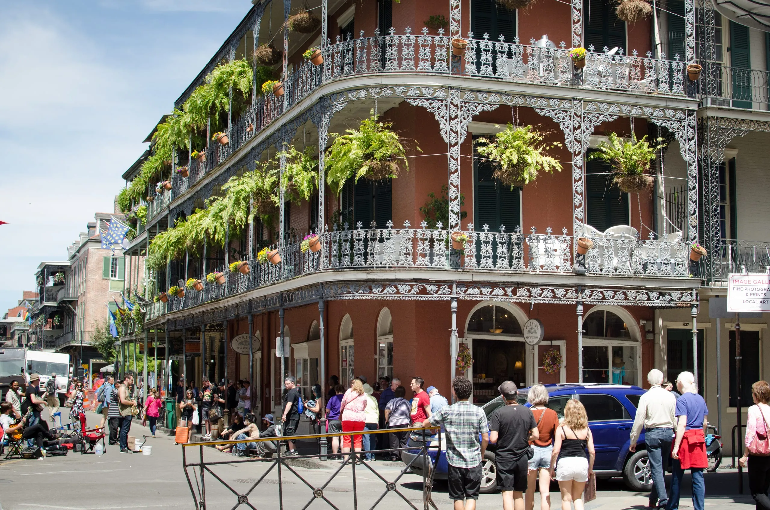 A French Quarter corner building draped in lacy cast-iron balconies and hanging ferns above a busy sidewalk crowd in New Orleans.