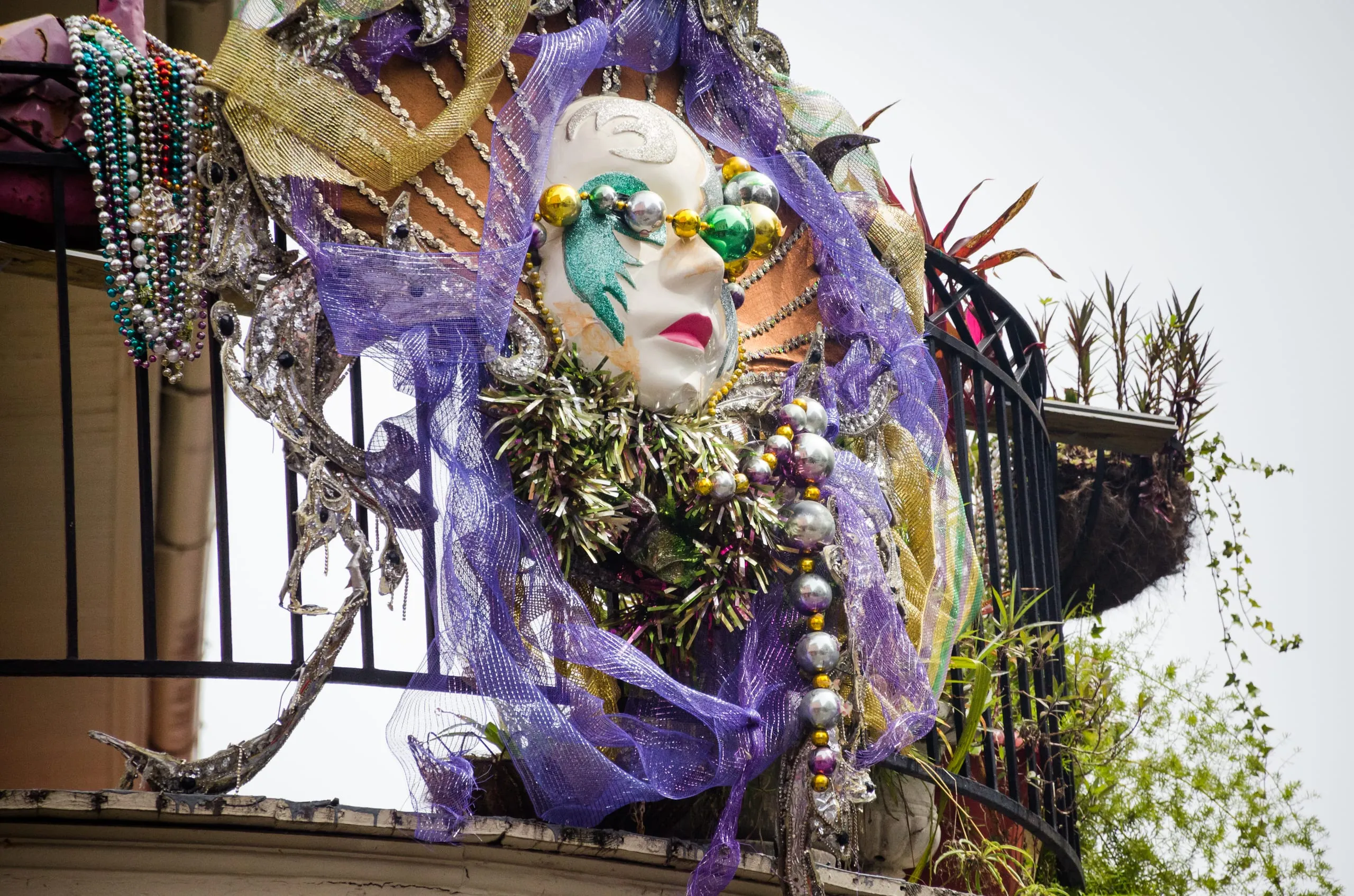 A white theatrical mask draped in purple mesh, beads, and tinsel decorating a wrought-iron balcony in New Orleans.
