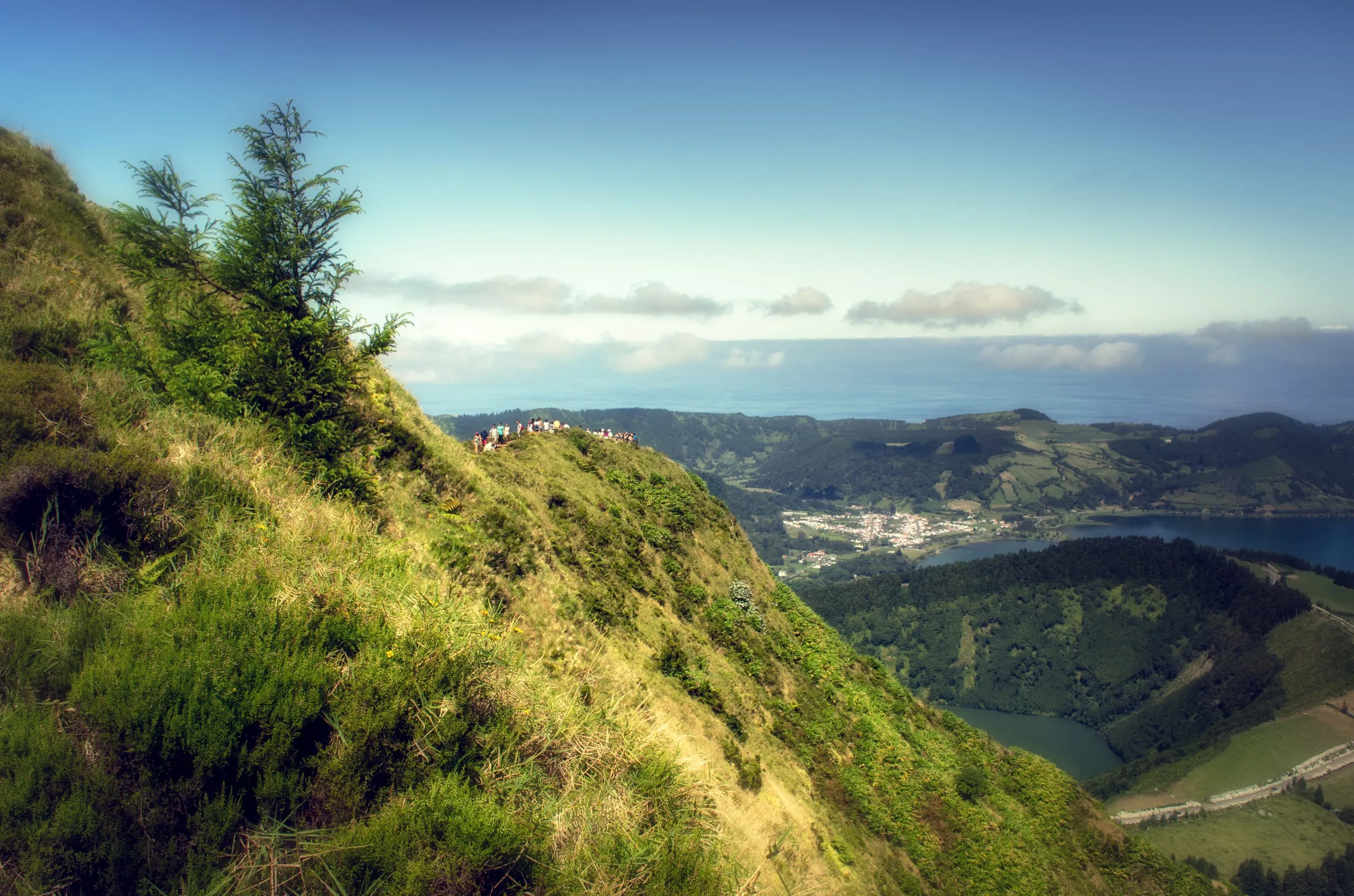 People stand on a grassy crater ridge overlooking a village, crater lake, and the ocean beyond under blue sky.