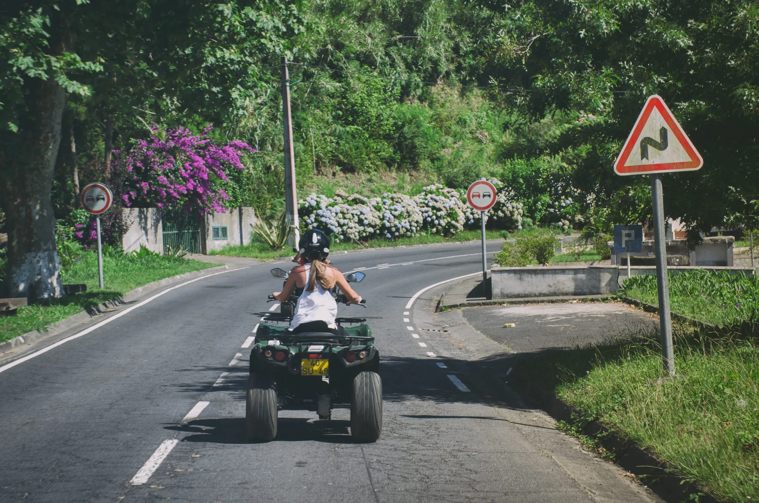 Two riders on a green quad bike drive down a curving country road past road signs and blue hydrangea hedges.