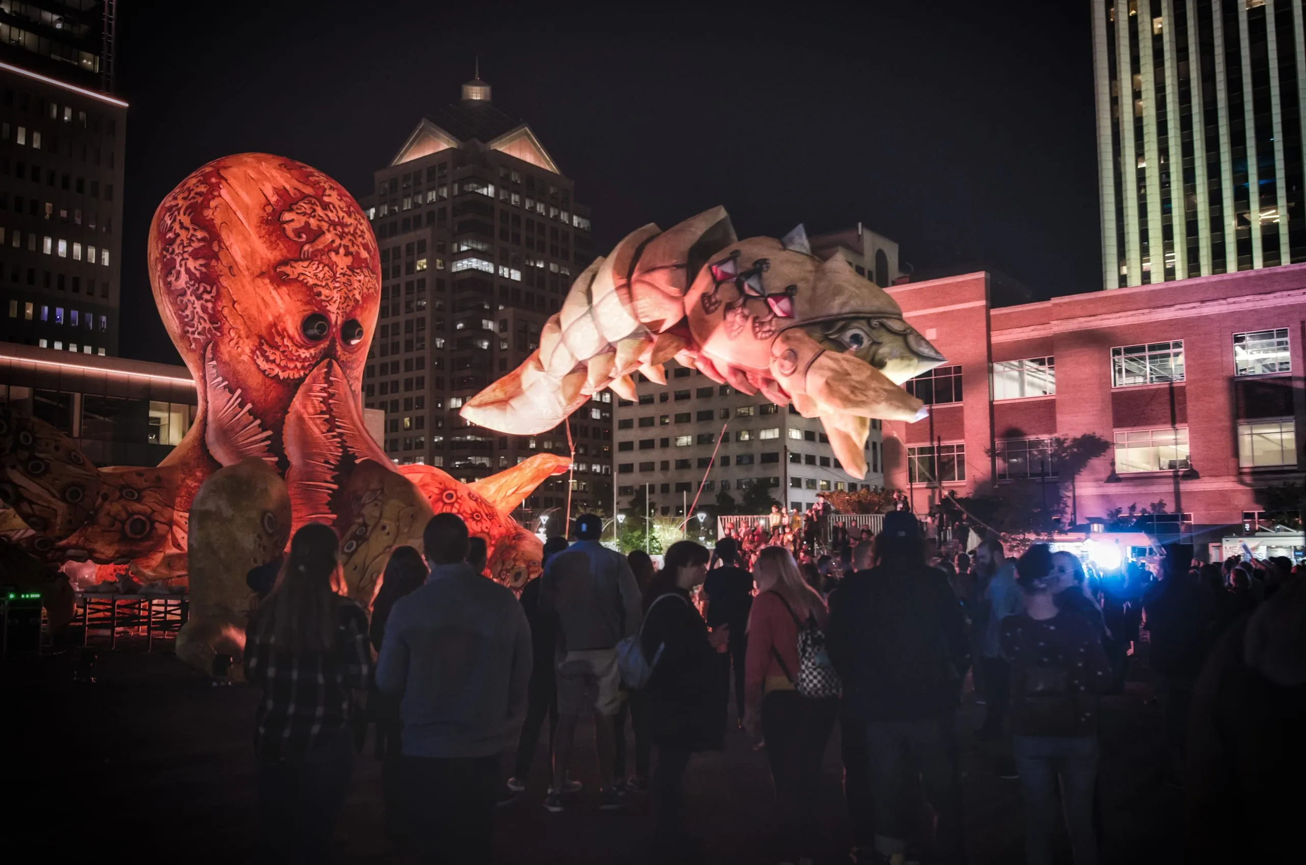 Giant illuminated octopus and crustacean puppets loom over a nighttime crowd amid downtown towers in Rochester, New York.