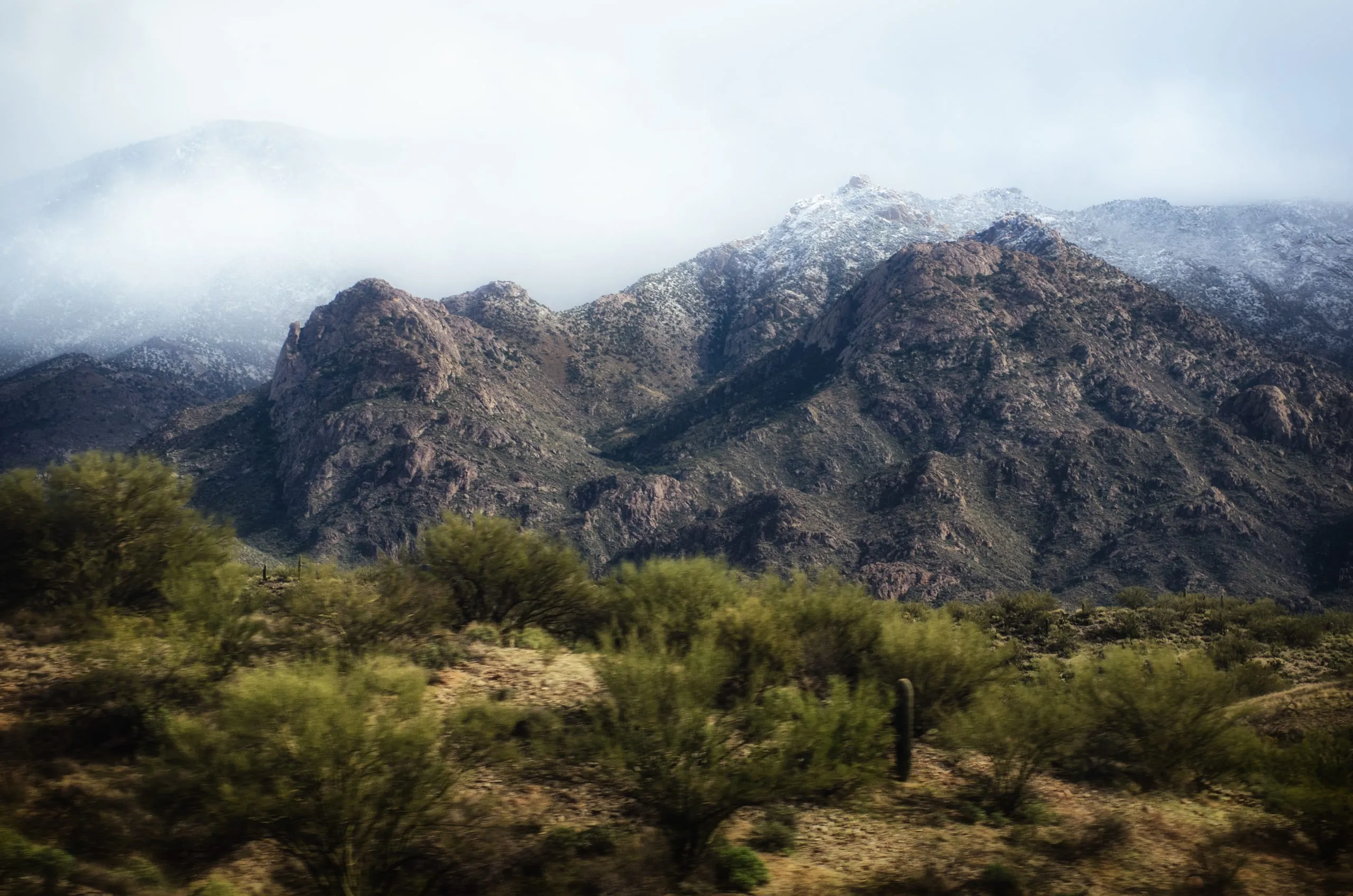 Snow lightly caps rugged desert mountains above green scrub and saguaro cactus in Arizona.