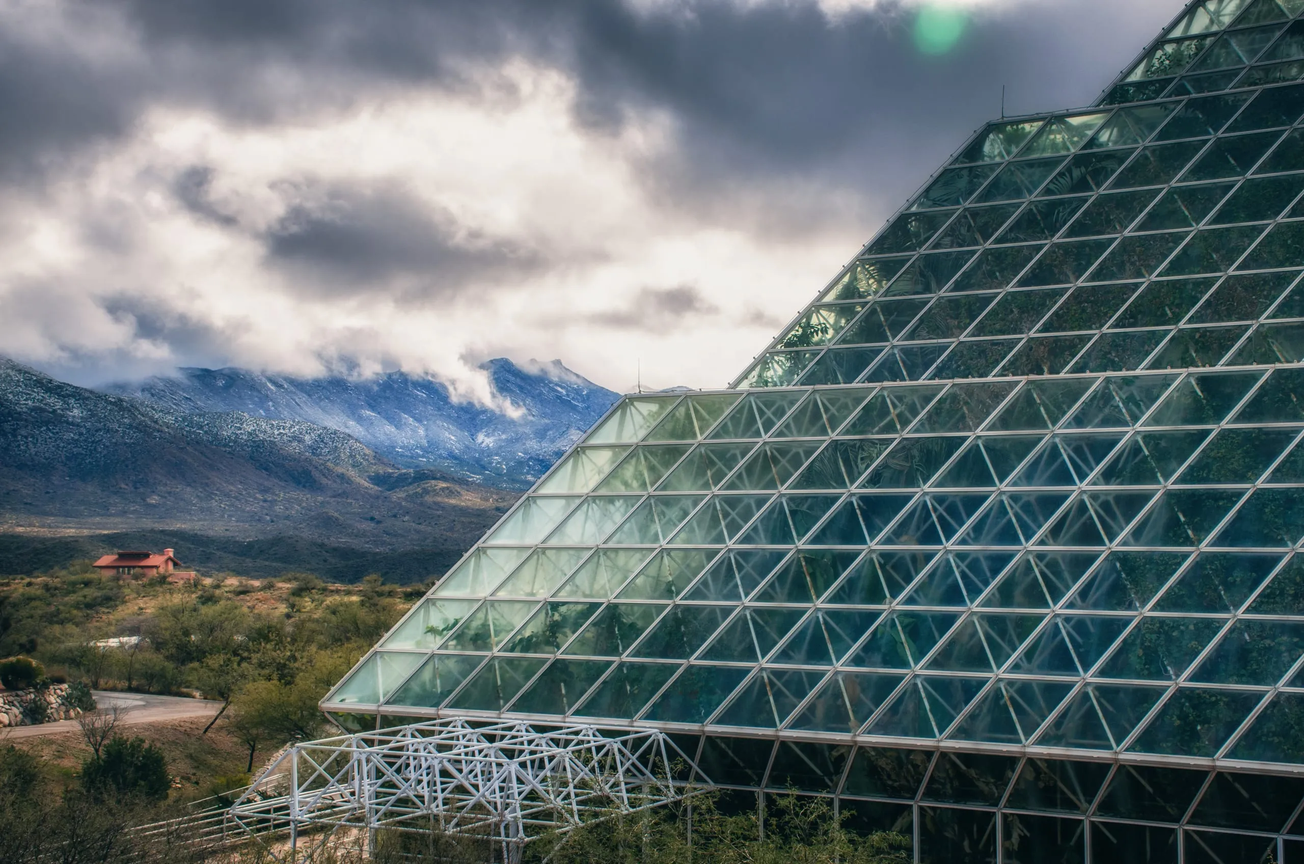 A vast triangular glass greenhouse rises against snow-dusted mountains and heavy clouds in Arizona.