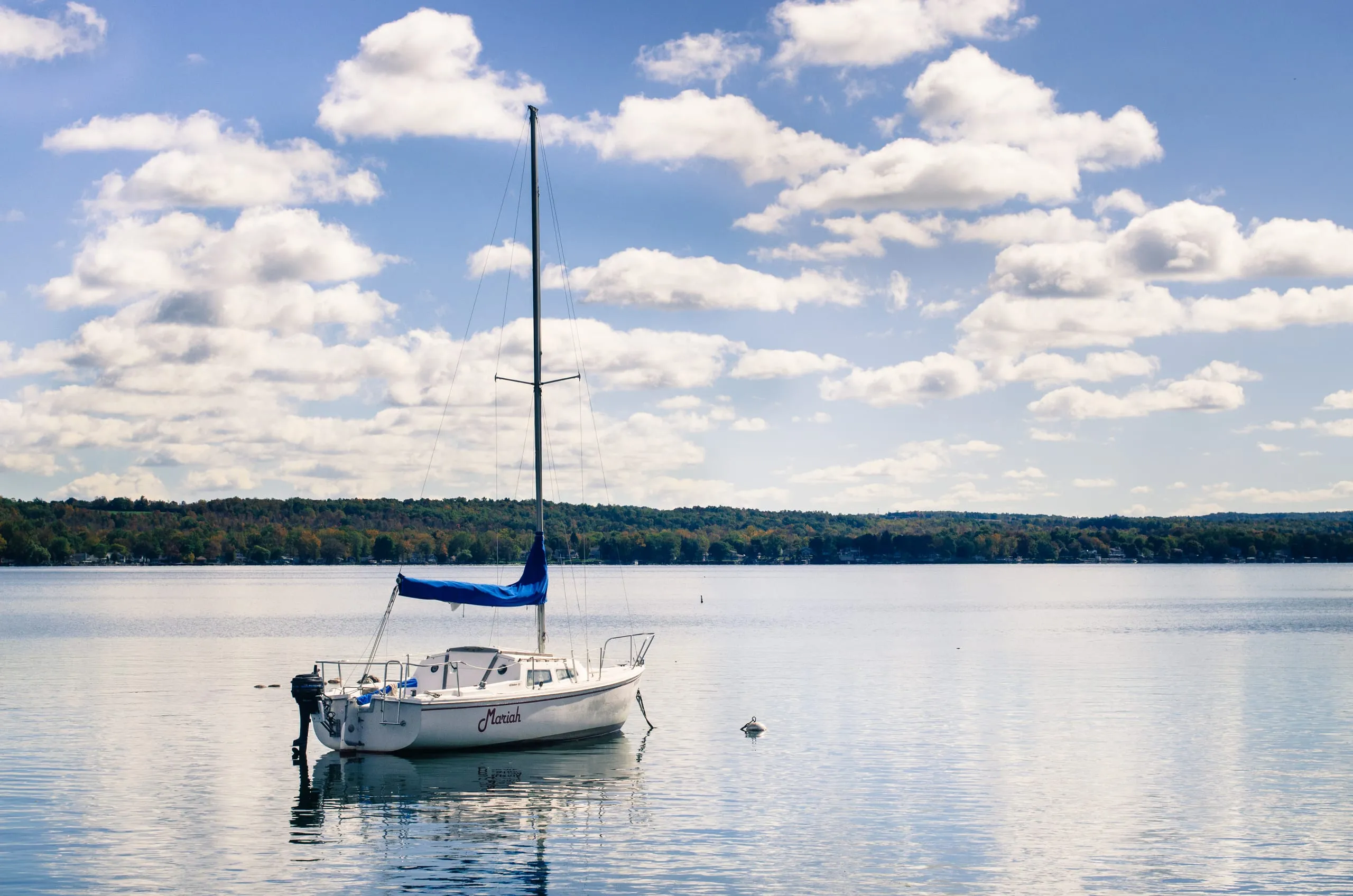 A white sailboat named Mariah with a blue sail cover moored on a calm Finger Lakes lake under puffy clouds.