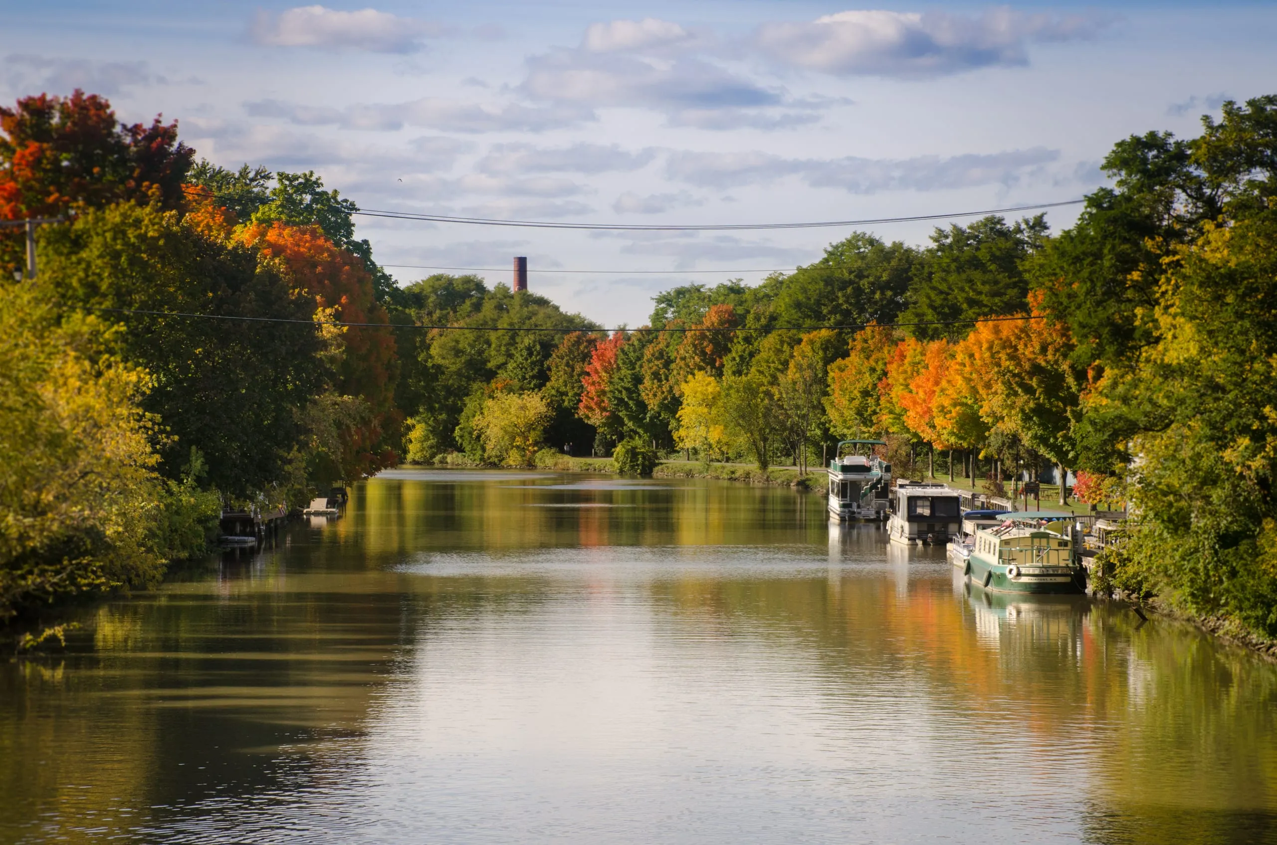 A still canal lined with turning fall foliage in the Finger Lakes, houseboats docked on the right and a brick smokestack beyond.